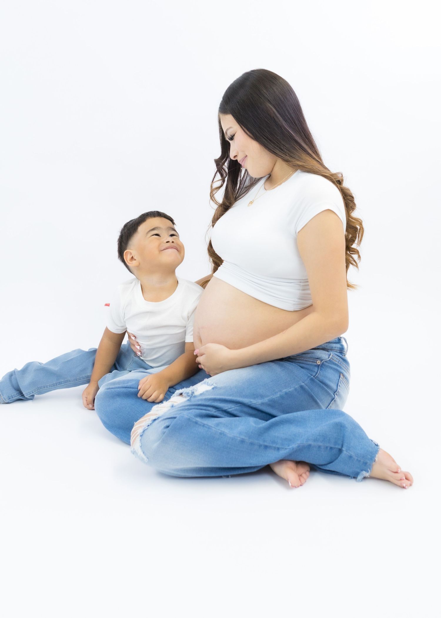 Pregnant woman in white crop top and jeans sits with young son on white background during maternity photo session.