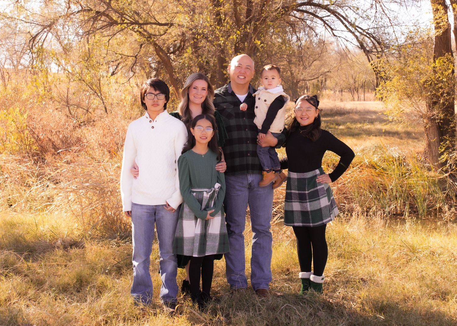 Family of six posing outdoors in autumn, surrounded by golden trees and fallen leaves in a scenic natural setting.