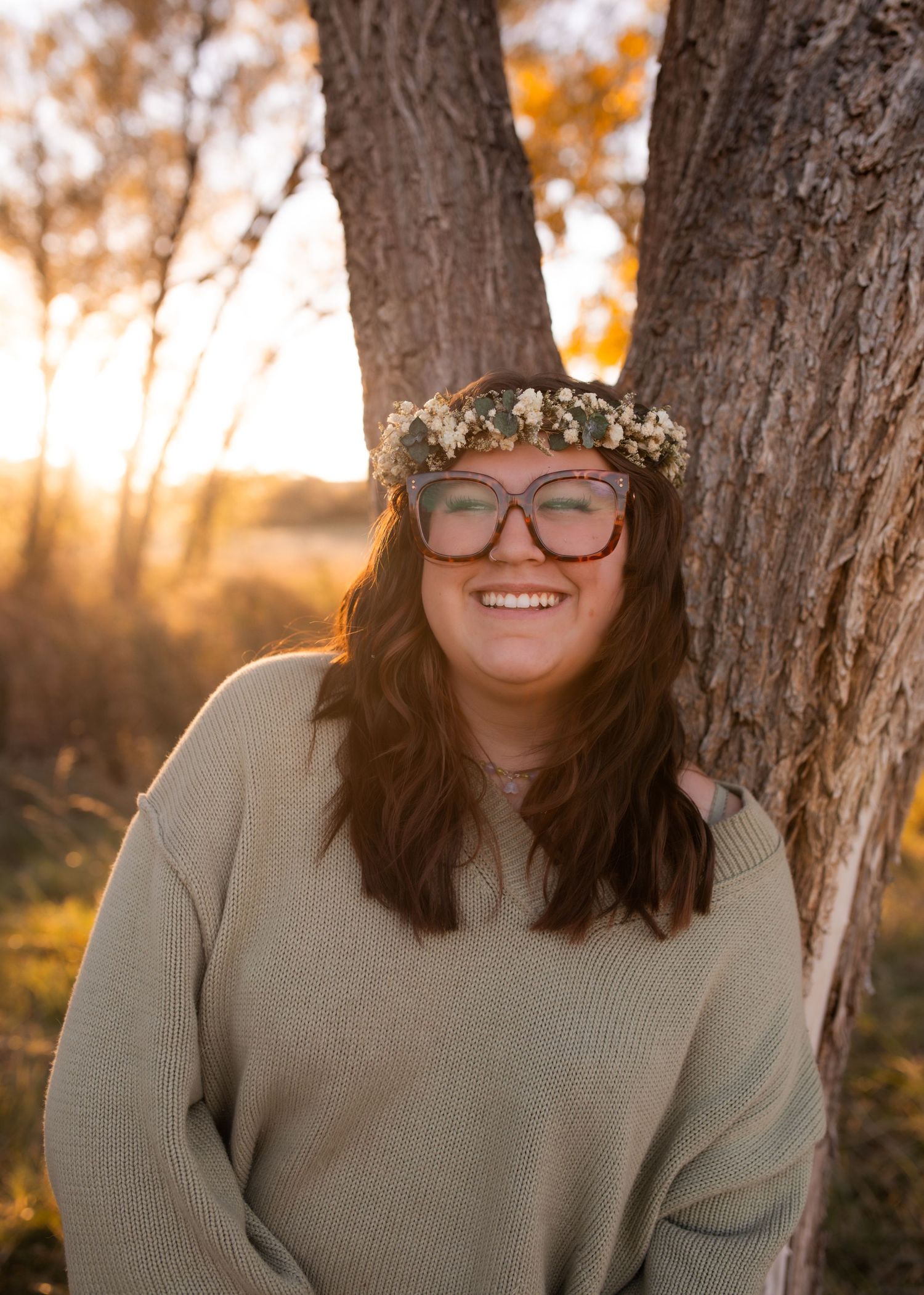 Smiling young woman with flower crown and red glasses leaning against tree at golden hour sunset.
