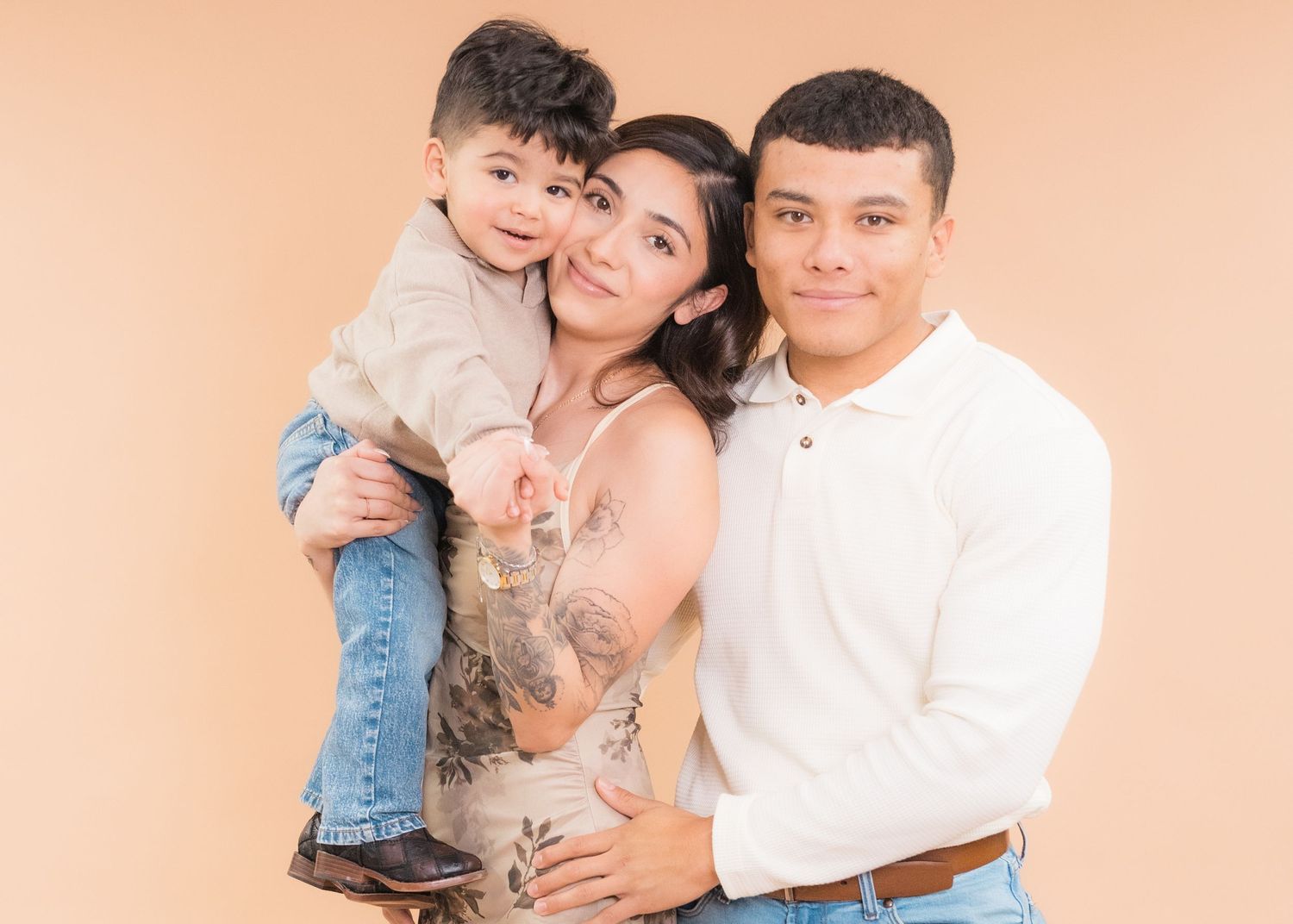 A young Hispanic family of three poses warmly against a peach background, mom holding toddler son beside tattooed dad.