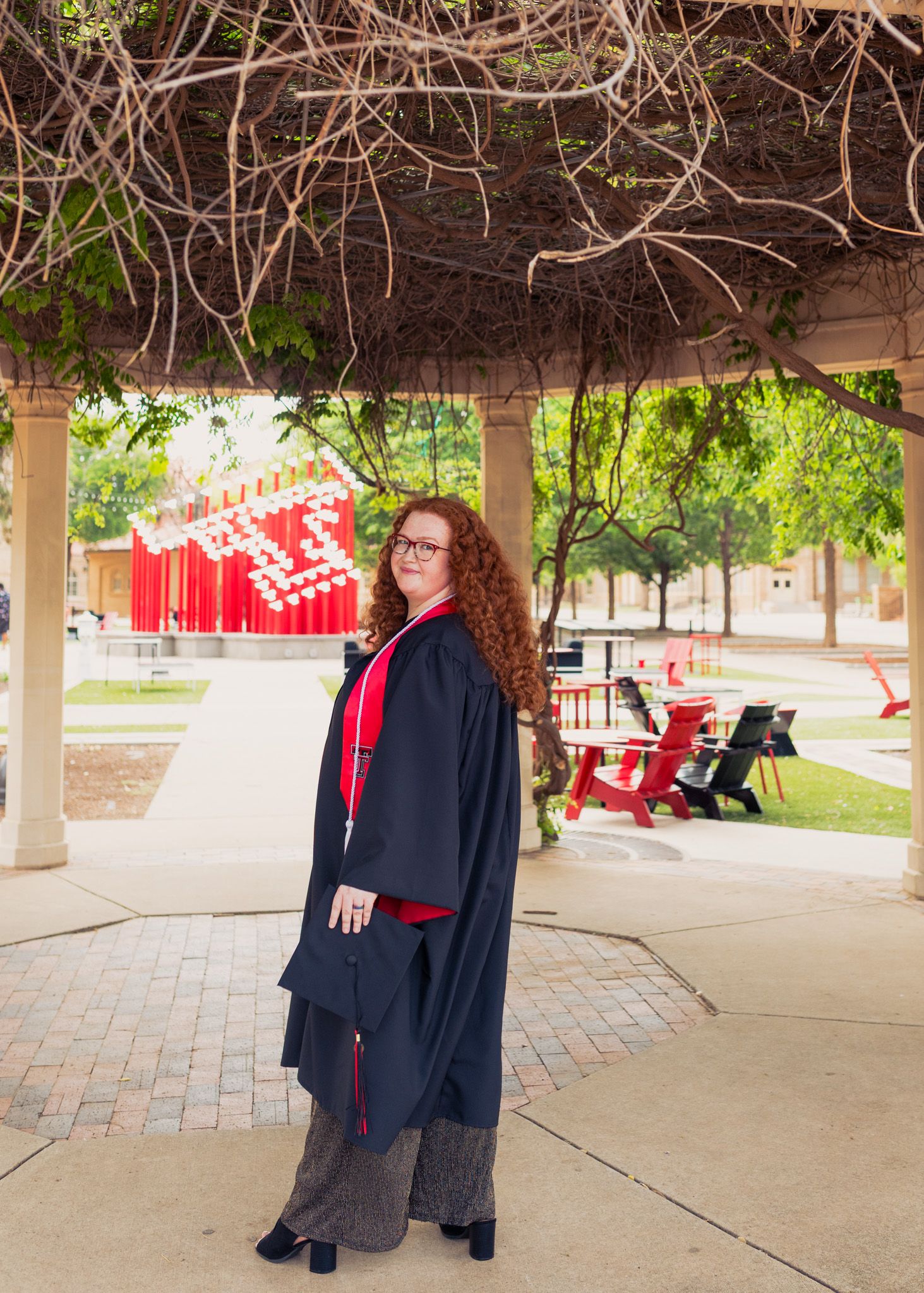 Graduate with red hair wearing traditional black gown stands under covered walkway on campus.
