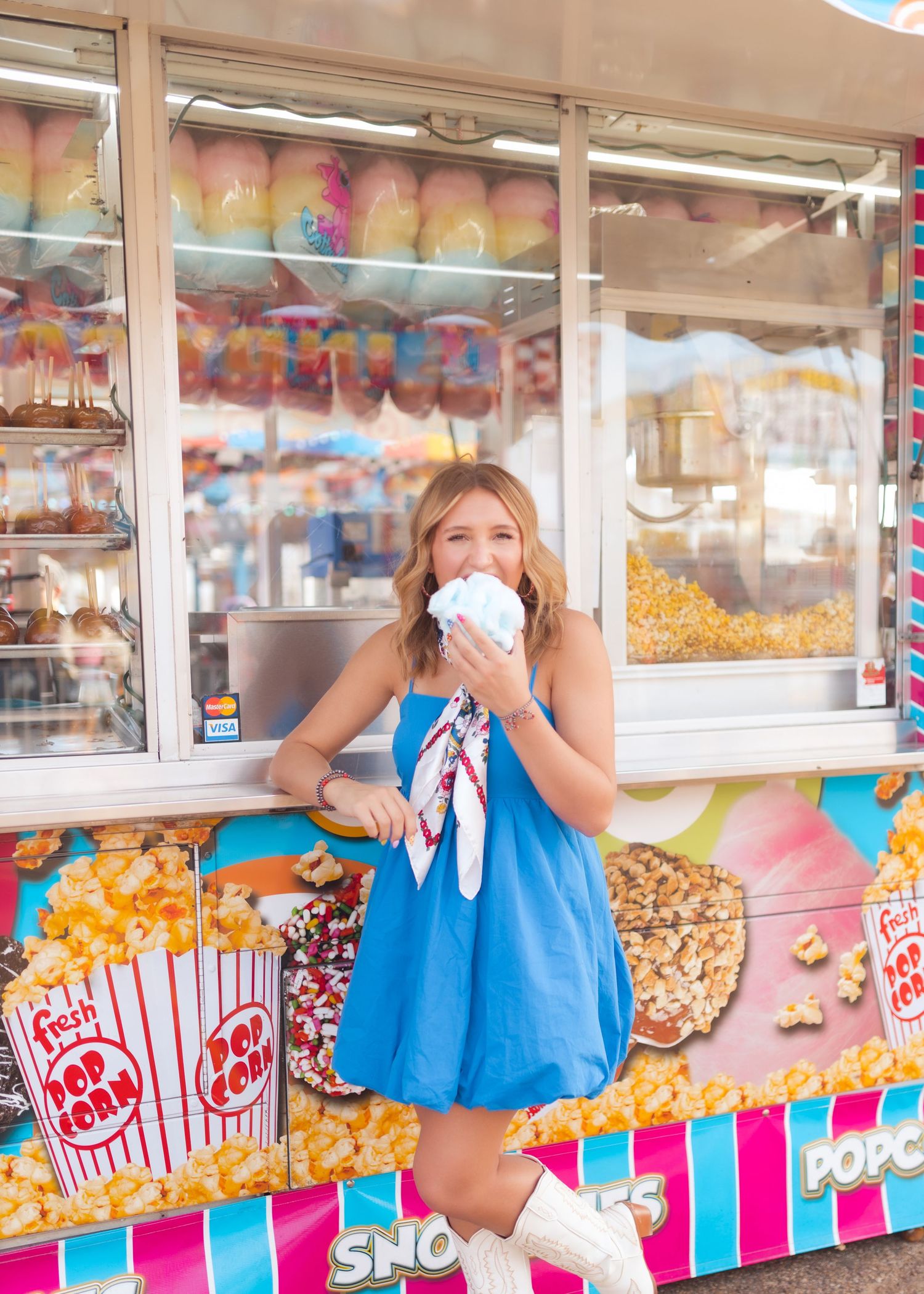 Young woman in blue dress eating cotton candy at a colorful carnival popcorn and snack booth.