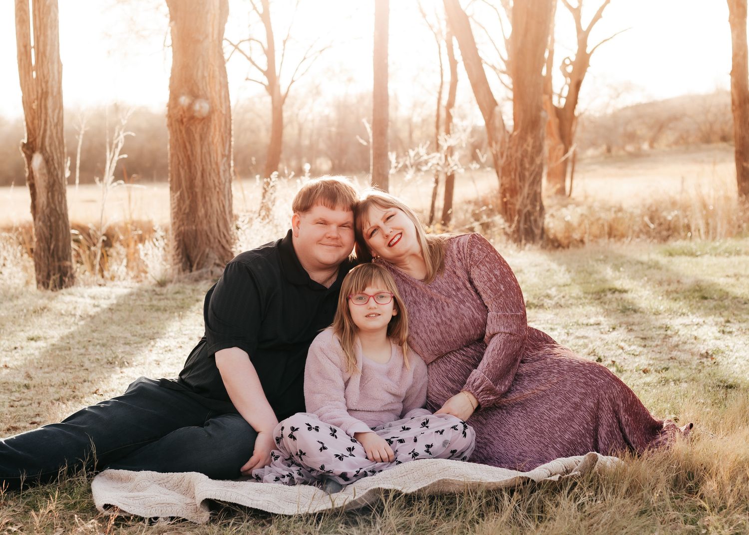 A warm family portrait shows three people sitting on a blanket outdoors in a serene natural setting at sunset.