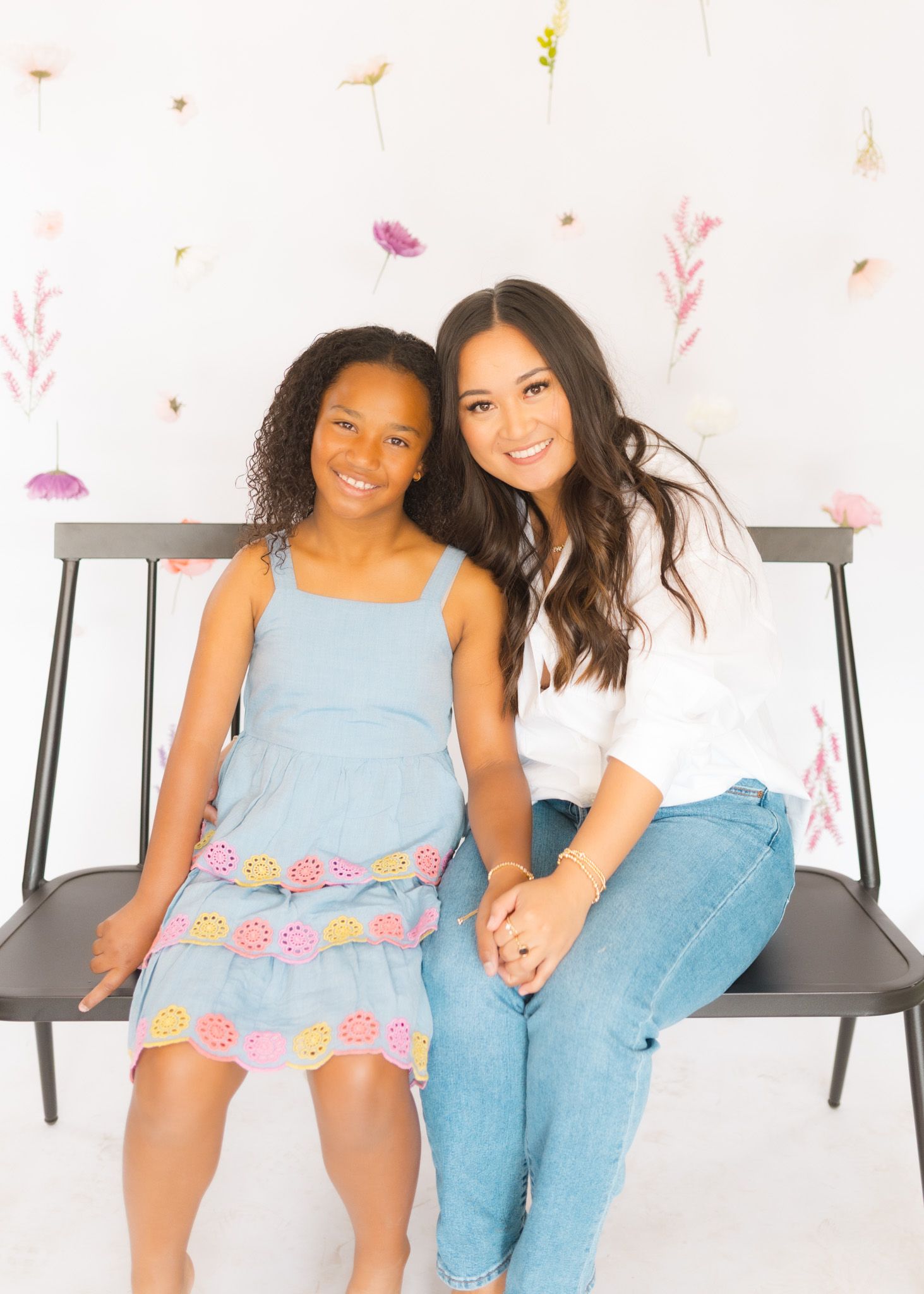 Two people sitting together on a bench against a floral backdrop in casual attire.