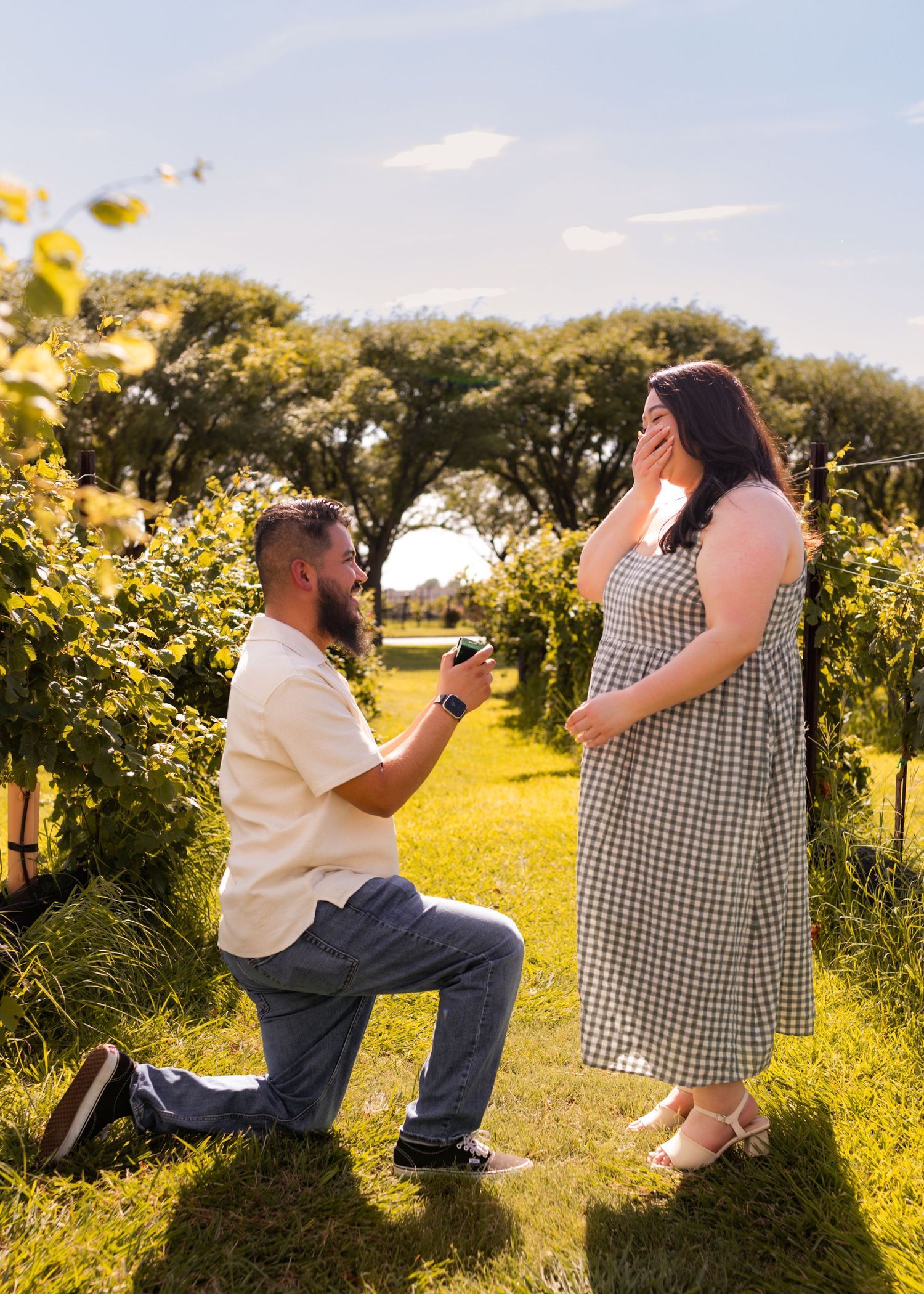 Man proposing on one knee to woman in checkered dress at a sunny vineyard, woman covers mouth in surprise.
