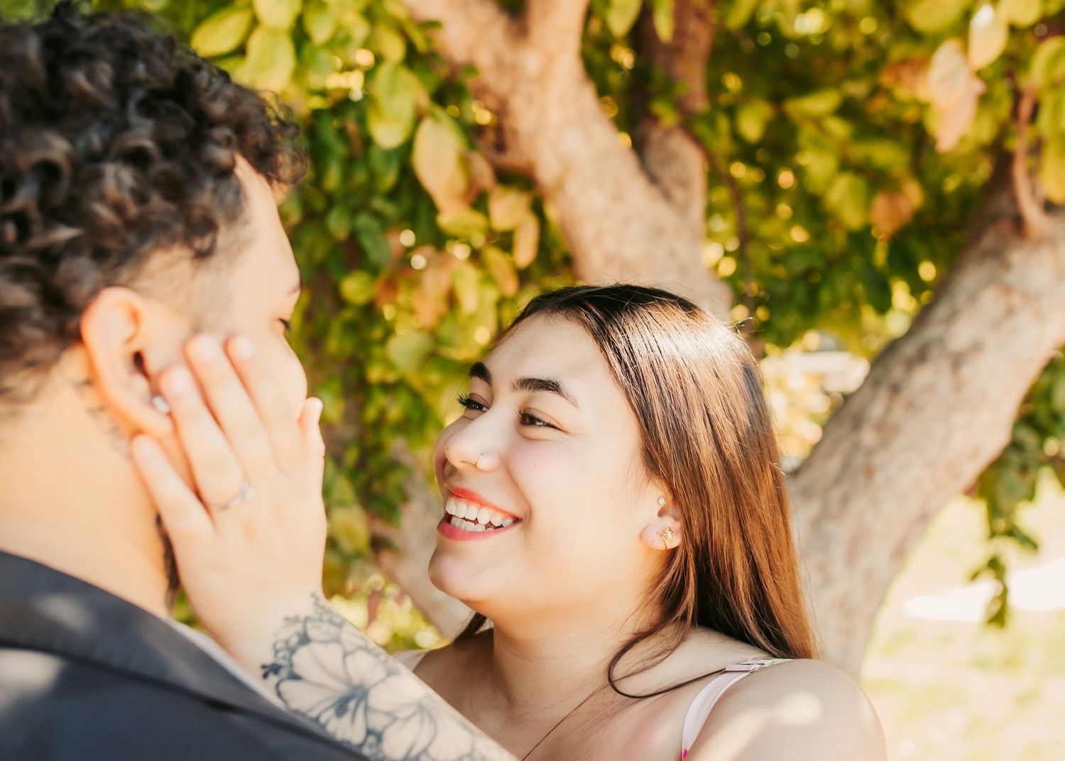 Two people share a sweet romantic moment smiling at each other in a park with green trees and sunlight.