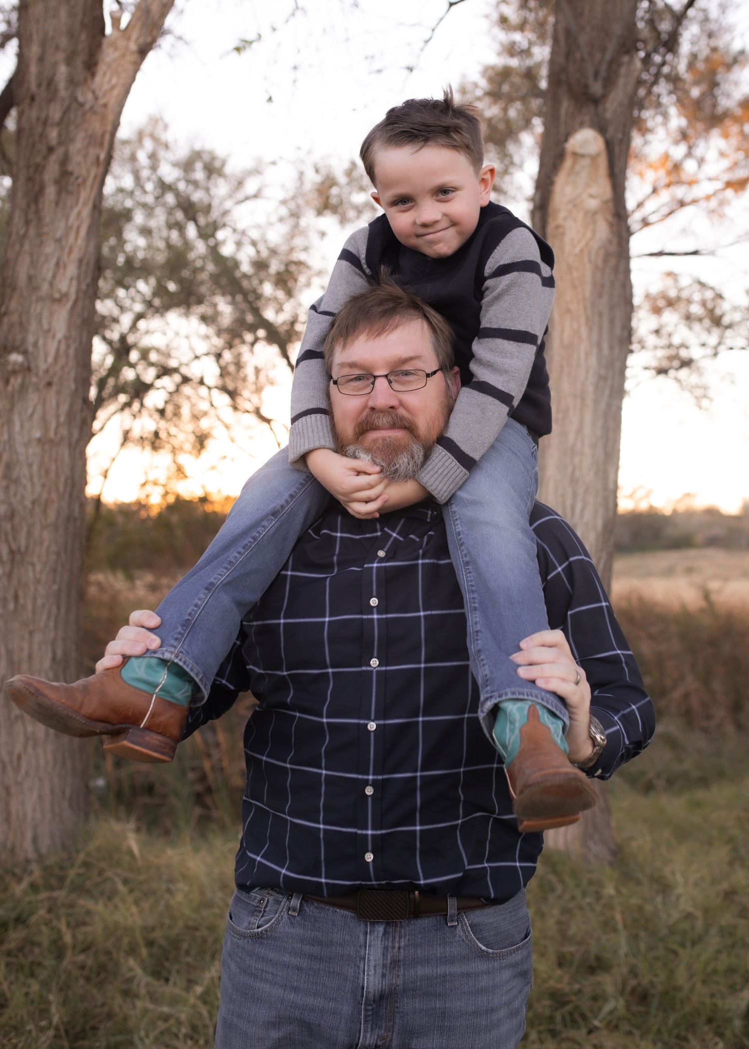 A man with glasses and a beard carries a smiling young boy on his shoulders outdoors among trees at sunset.