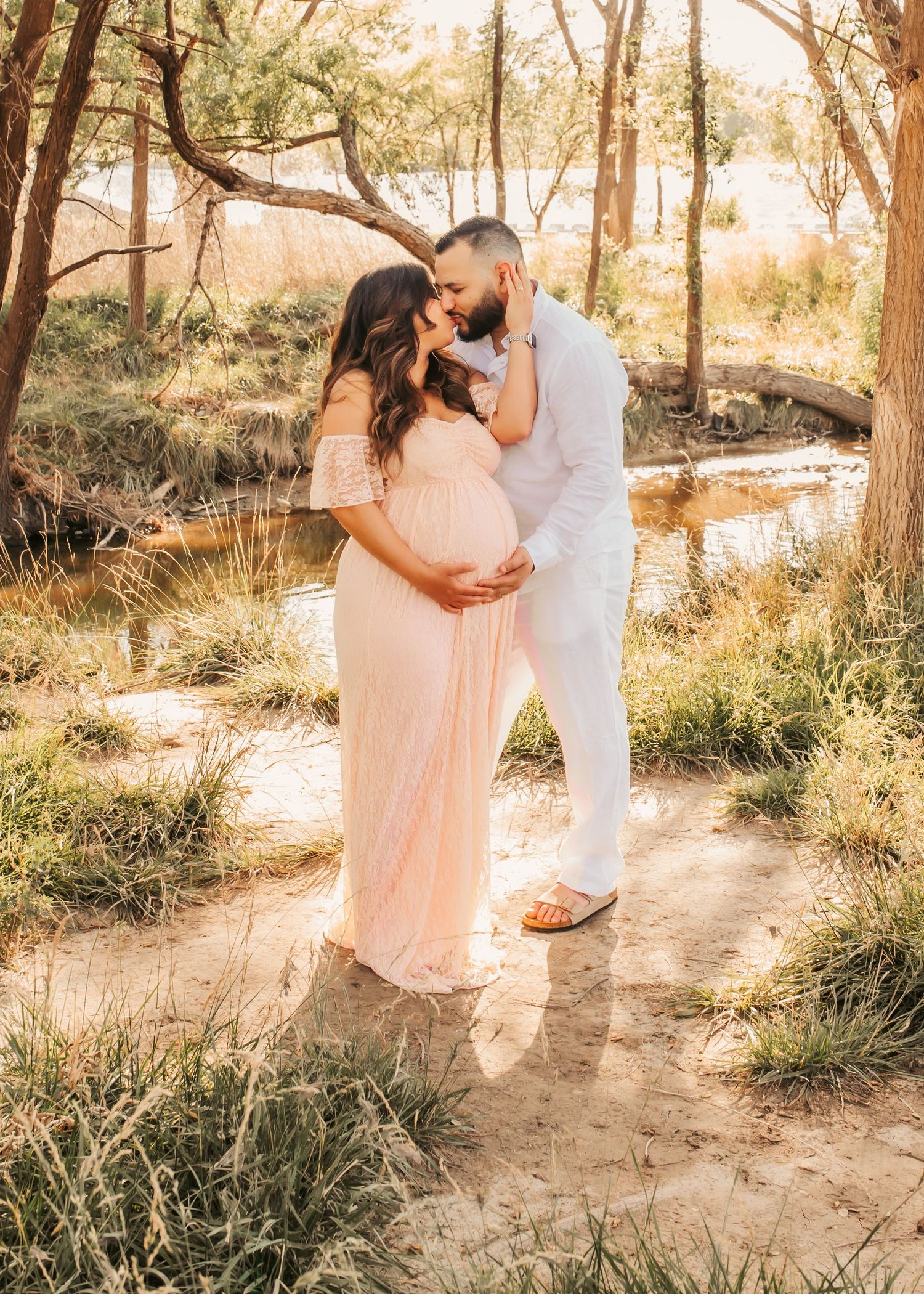 Couple sharing a tender kiss outdoors, pregnant woman in blush maxi dress, man in white, surrounded by trees and creek.