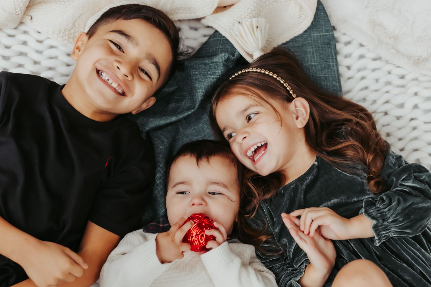 Three children lying on white blanket making silly faces and laughing together.