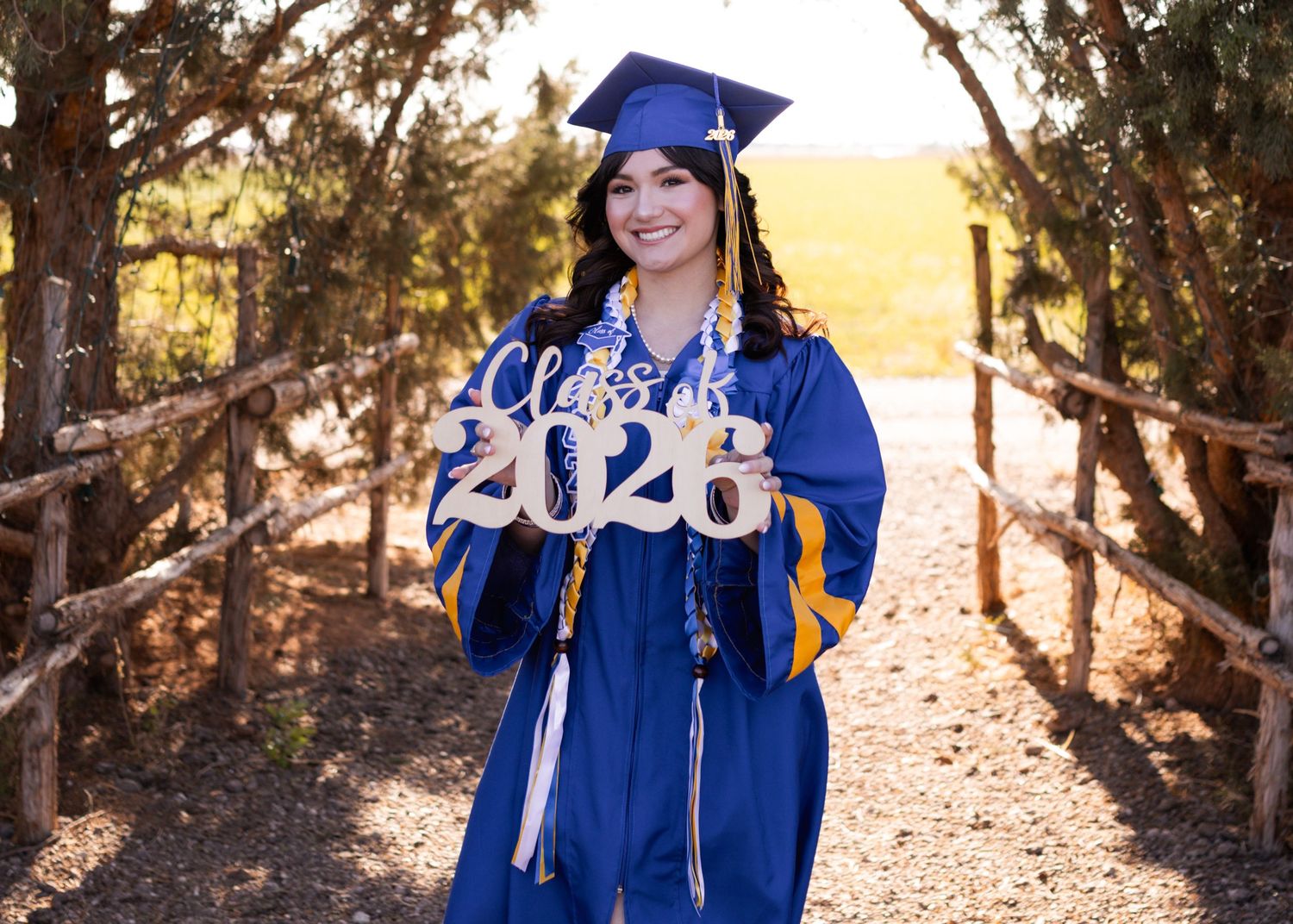 A smiling graduate in blue cap and gown holds a Class of 2026 sign outdoors on a sunny day.