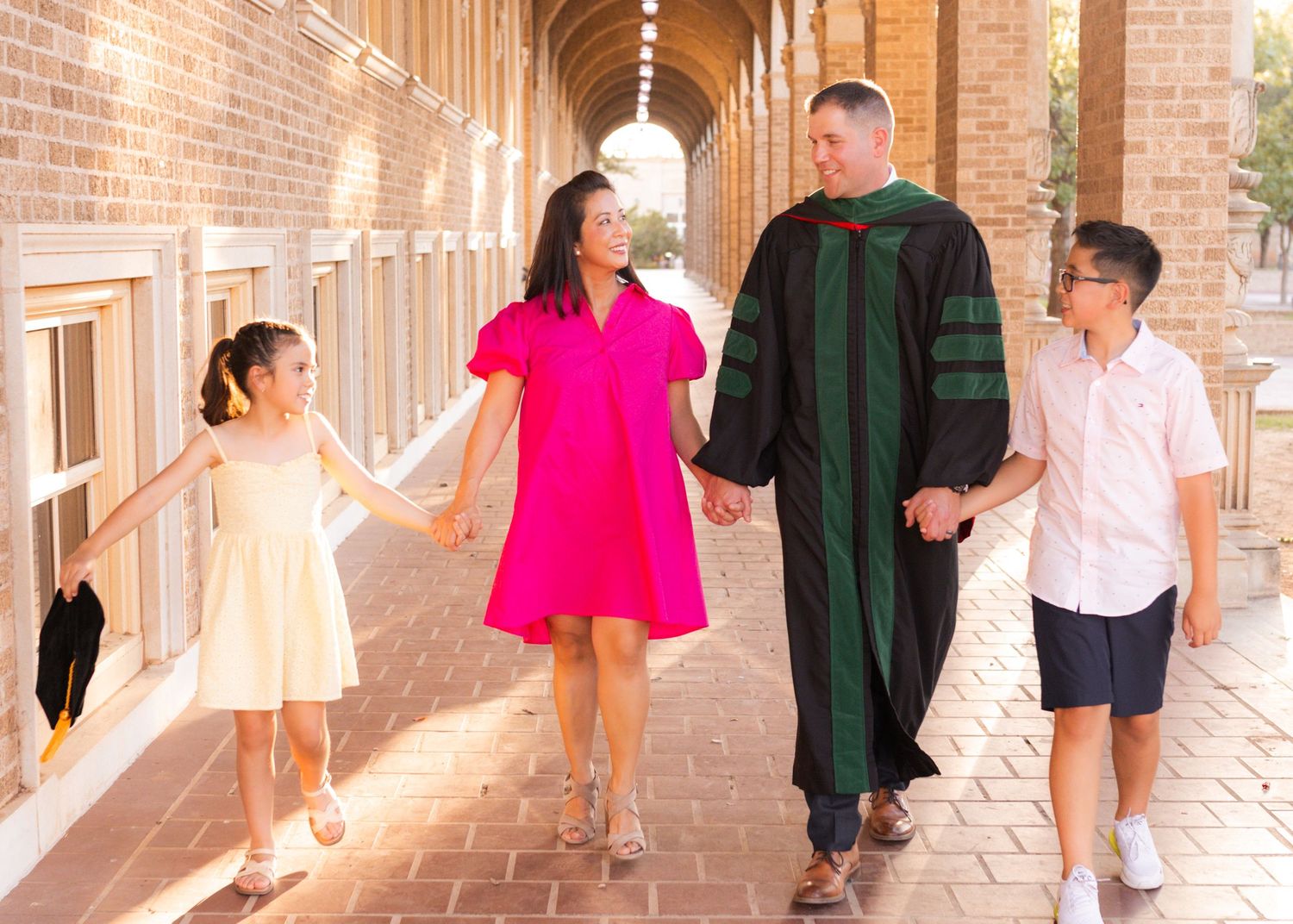 Family of four walks together under brick archway, man wearing green doctoral graduation gown, celebrating academic achievement.