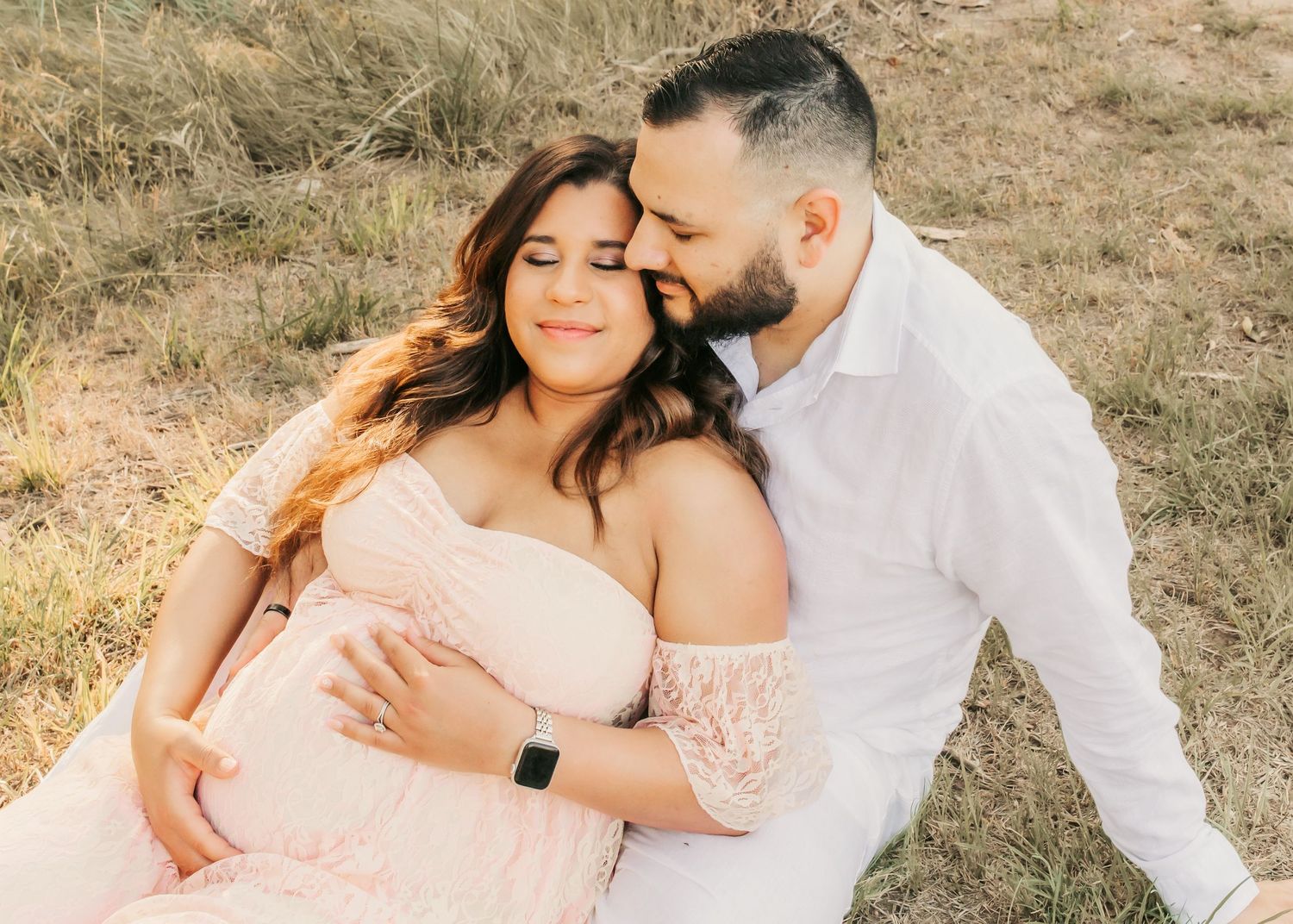 Couple shares tender maternity photo outdoors, woman in pink lace dress cradling baby bump while man kisses her forehead.