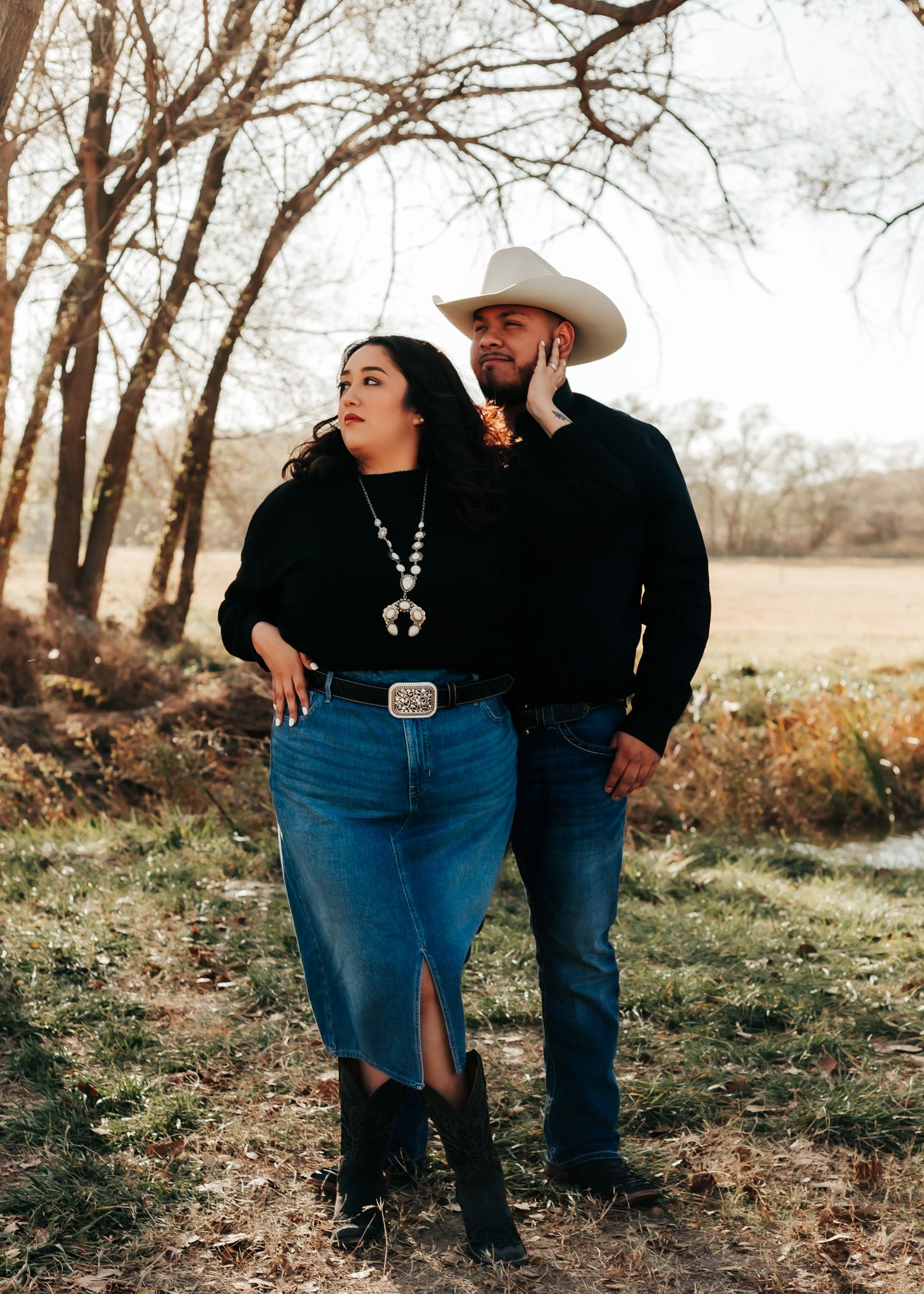 Couple in western attire posing together outdoors with trees and sunlight in background.