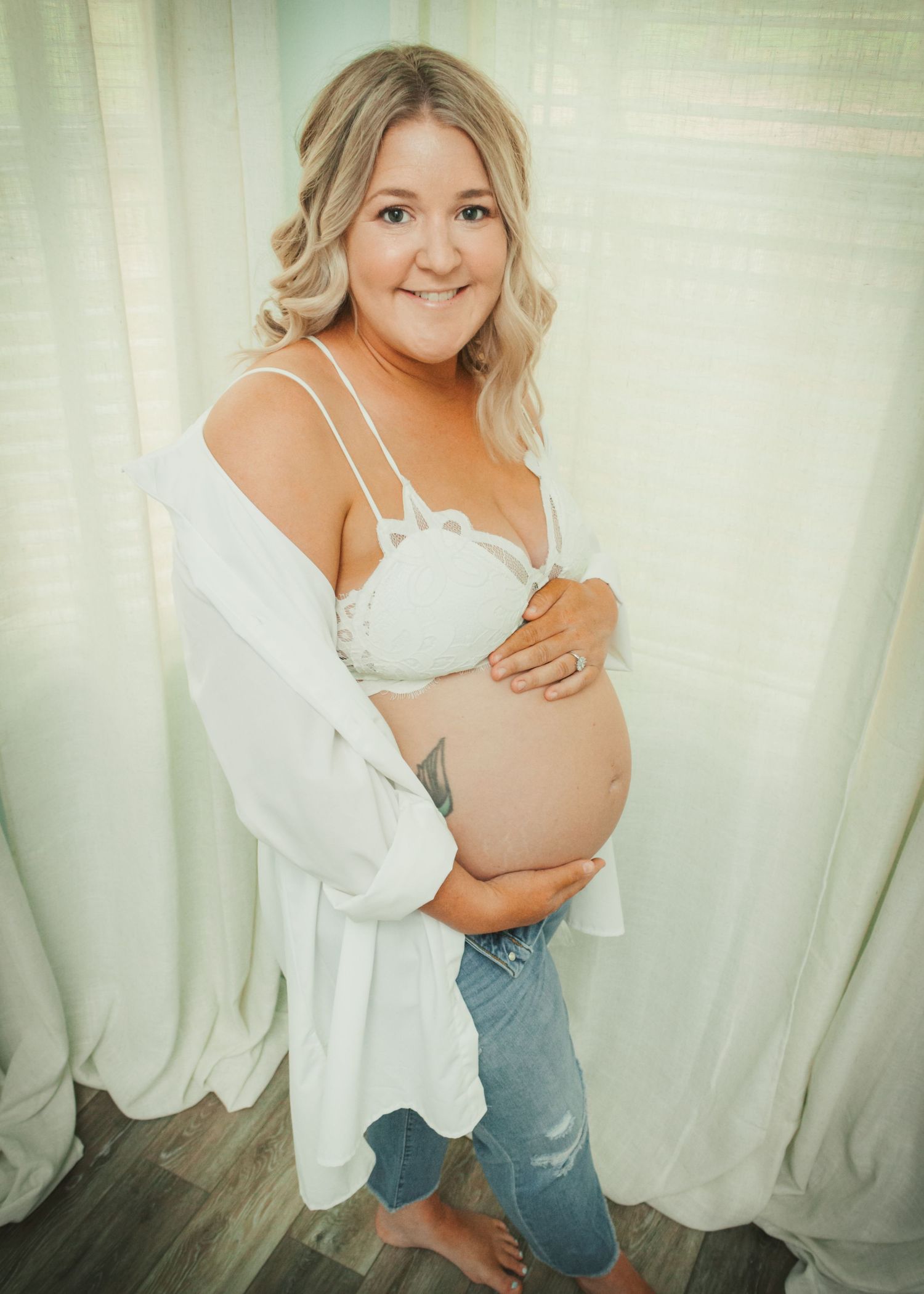 Indoor maternity photo against white curtains wearing white robe and jeans.