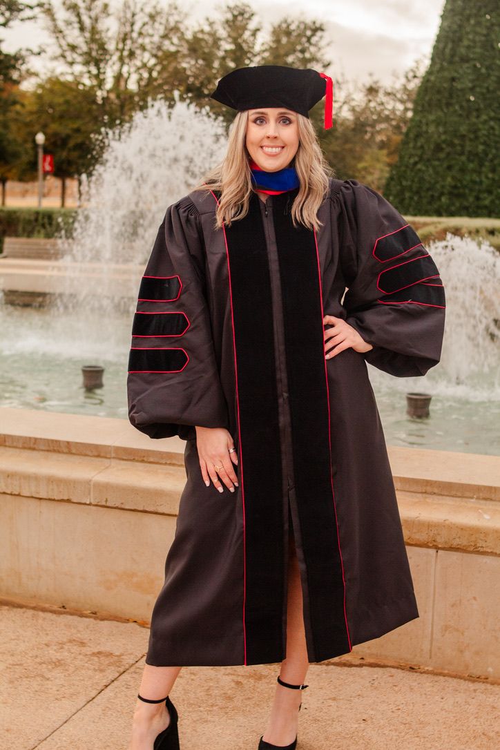 Doctoral graduate in black academic regalia with red trim poses by fountain in graduation photos.
