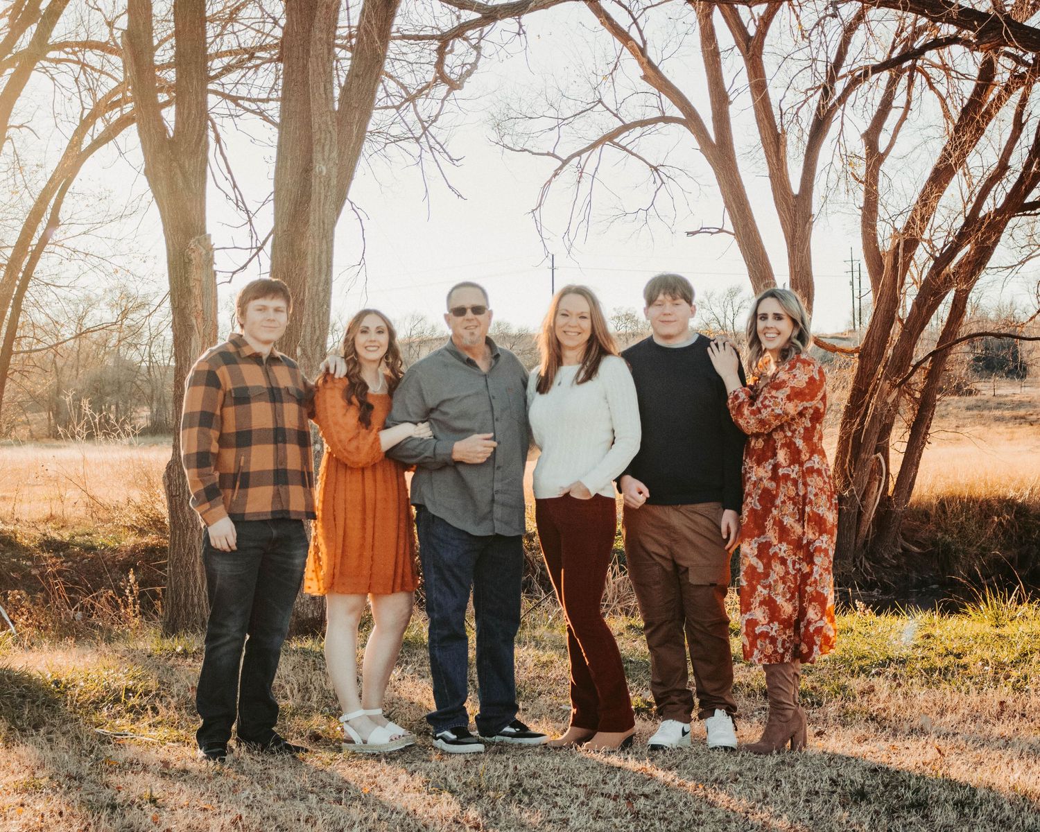 Group of people in casual fall clothing posed together outdoors among bare trees at sunset.