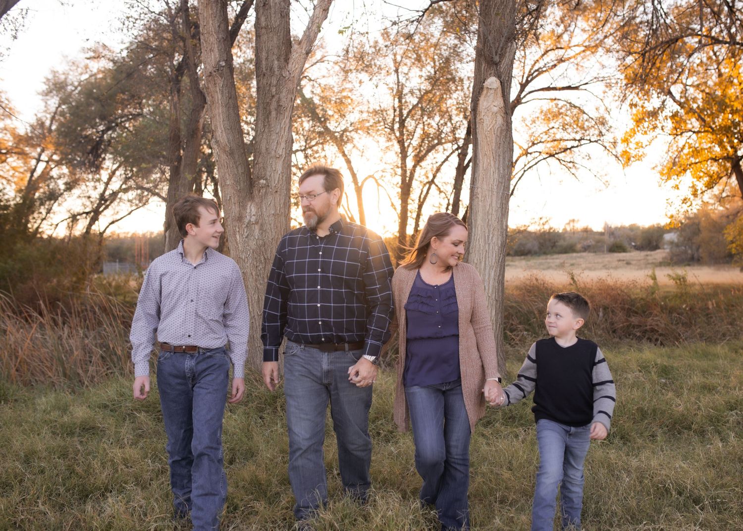 Family of four walking together in a golden autumn field with tall trees and warm sunset light in the background.