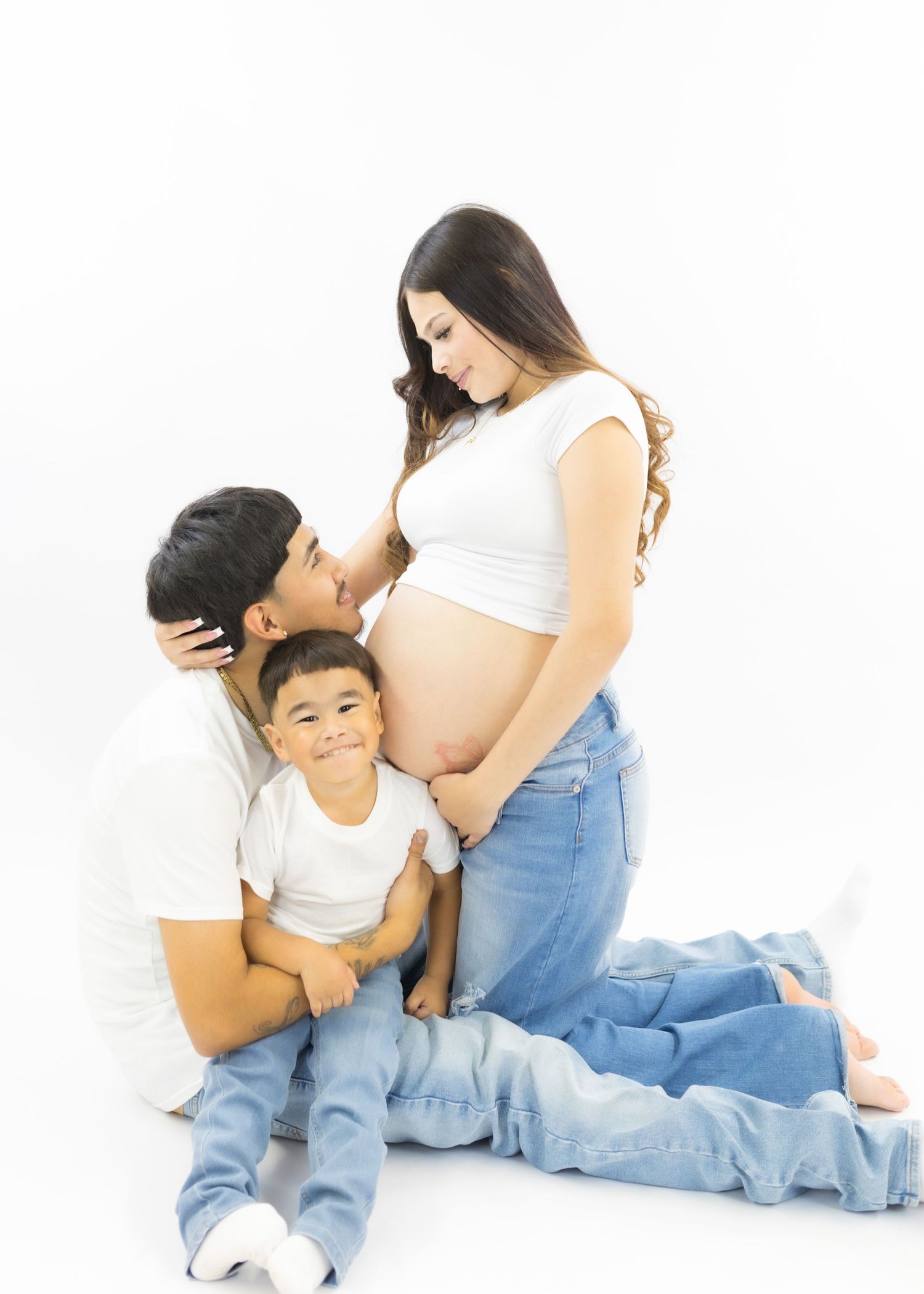 A pregnant woman in white crop top and jeans poses with her partner and toddler son in a bright white studio maternity photo.