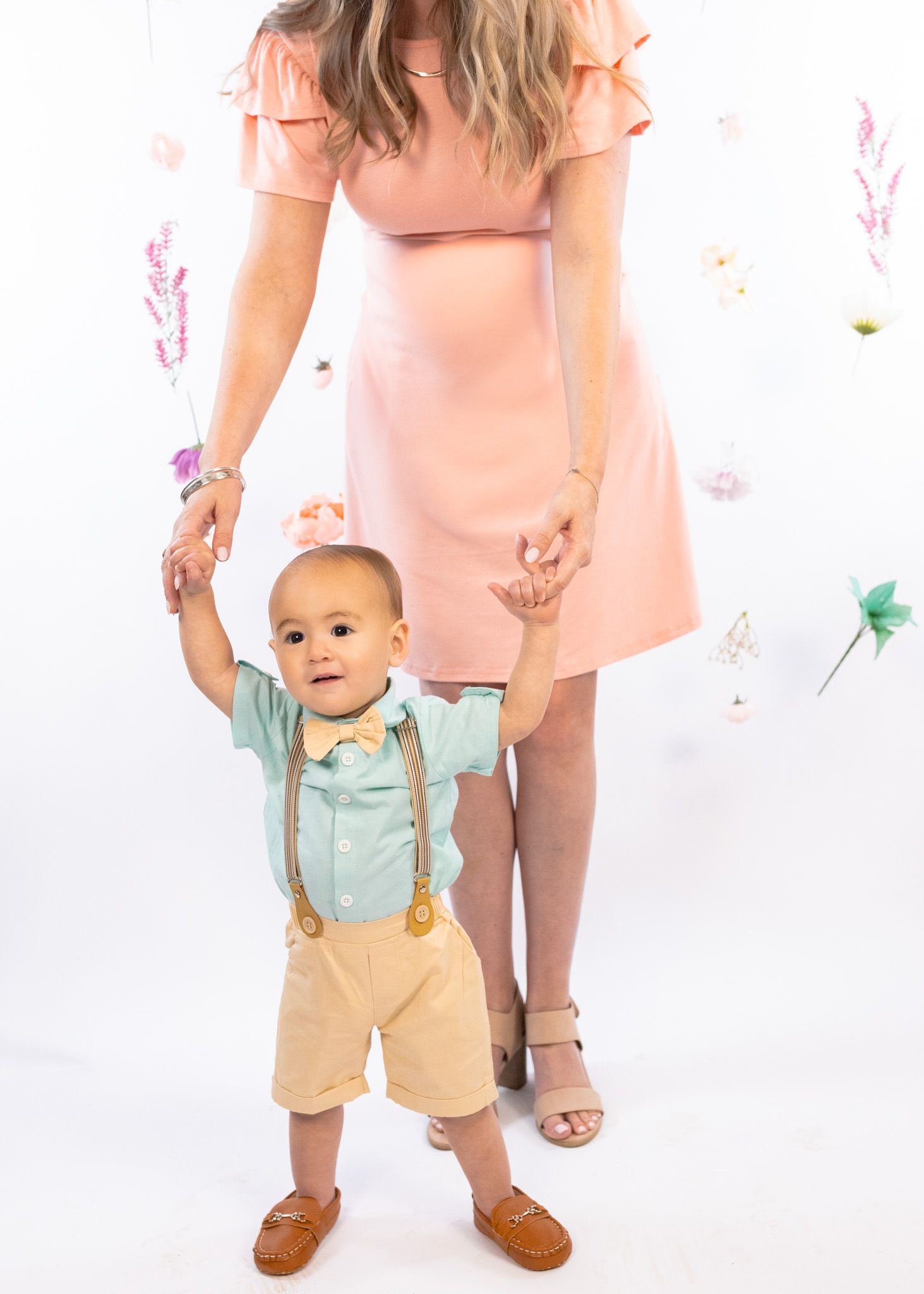 Parent and toddler in coordinating outfits pose against floral backdrop in studio setting.