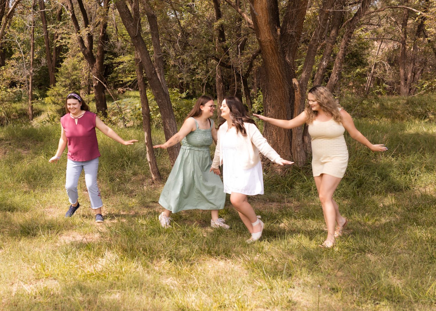 Four women in colorful dresses laugh and dance together in a sunny green meadow surrounded by trees.