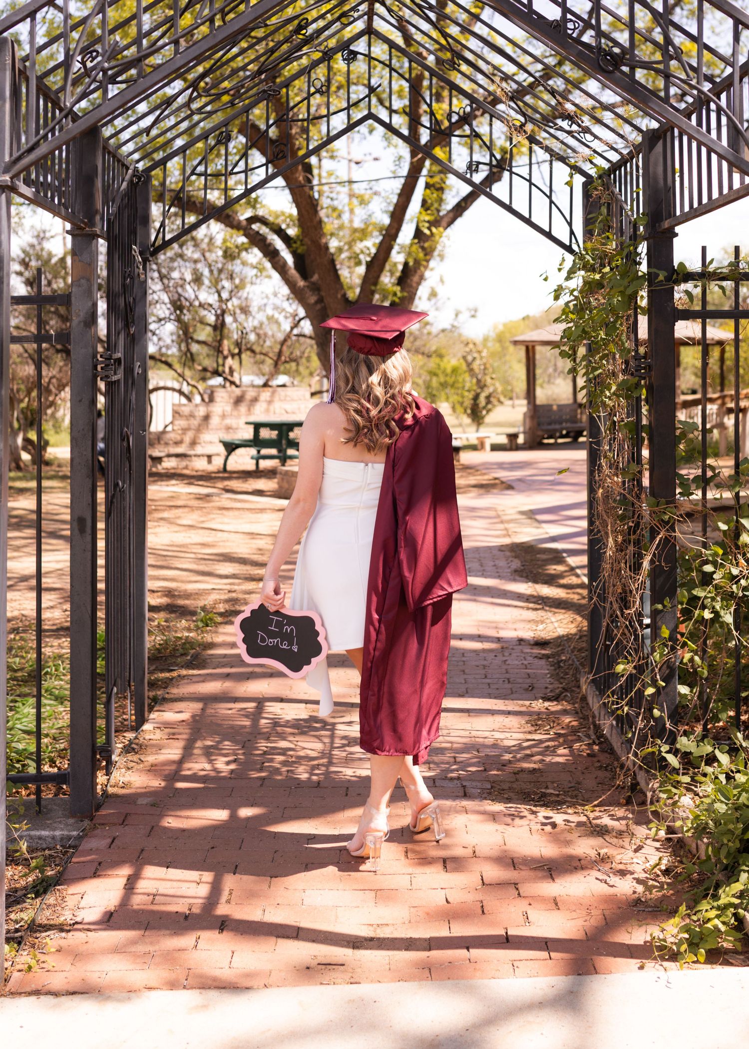 A graduate in a maroon cap and gown walks through an iron garden archway holding a chalkboard sign on a brick path.