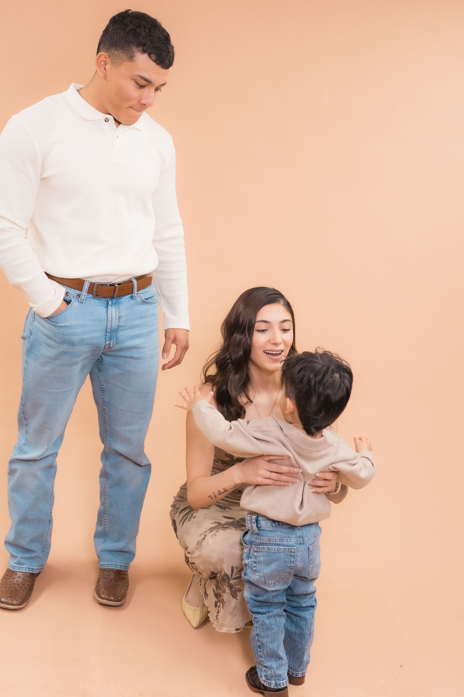 A young family with a mother hugging her son while the father looks on, all wearing casual jeans outfits on a beige background.