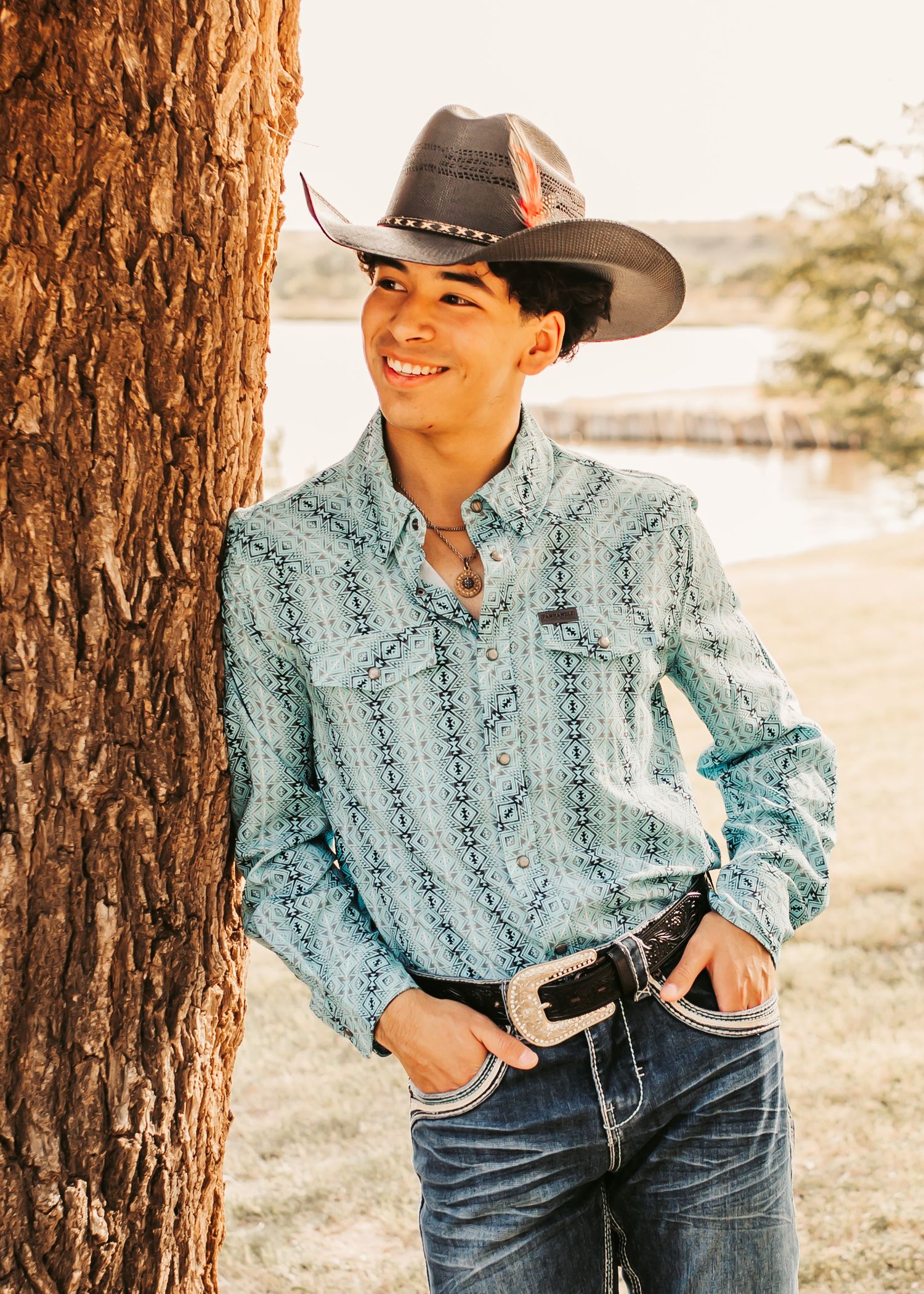 Cowboy in blue patterned shirt and hat leaning against tree trunk.