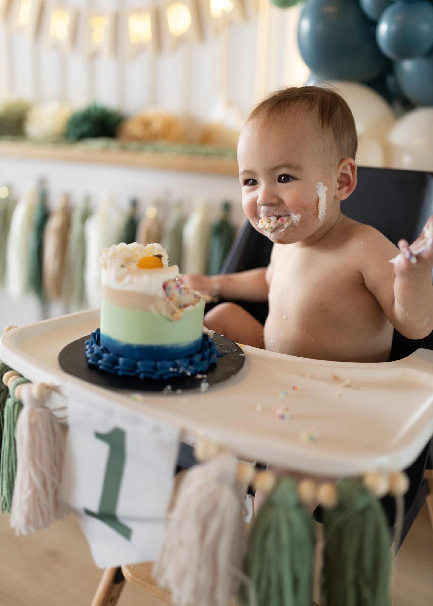 Messy baby celebrates first birthday with blue and green smash cake on highchair.