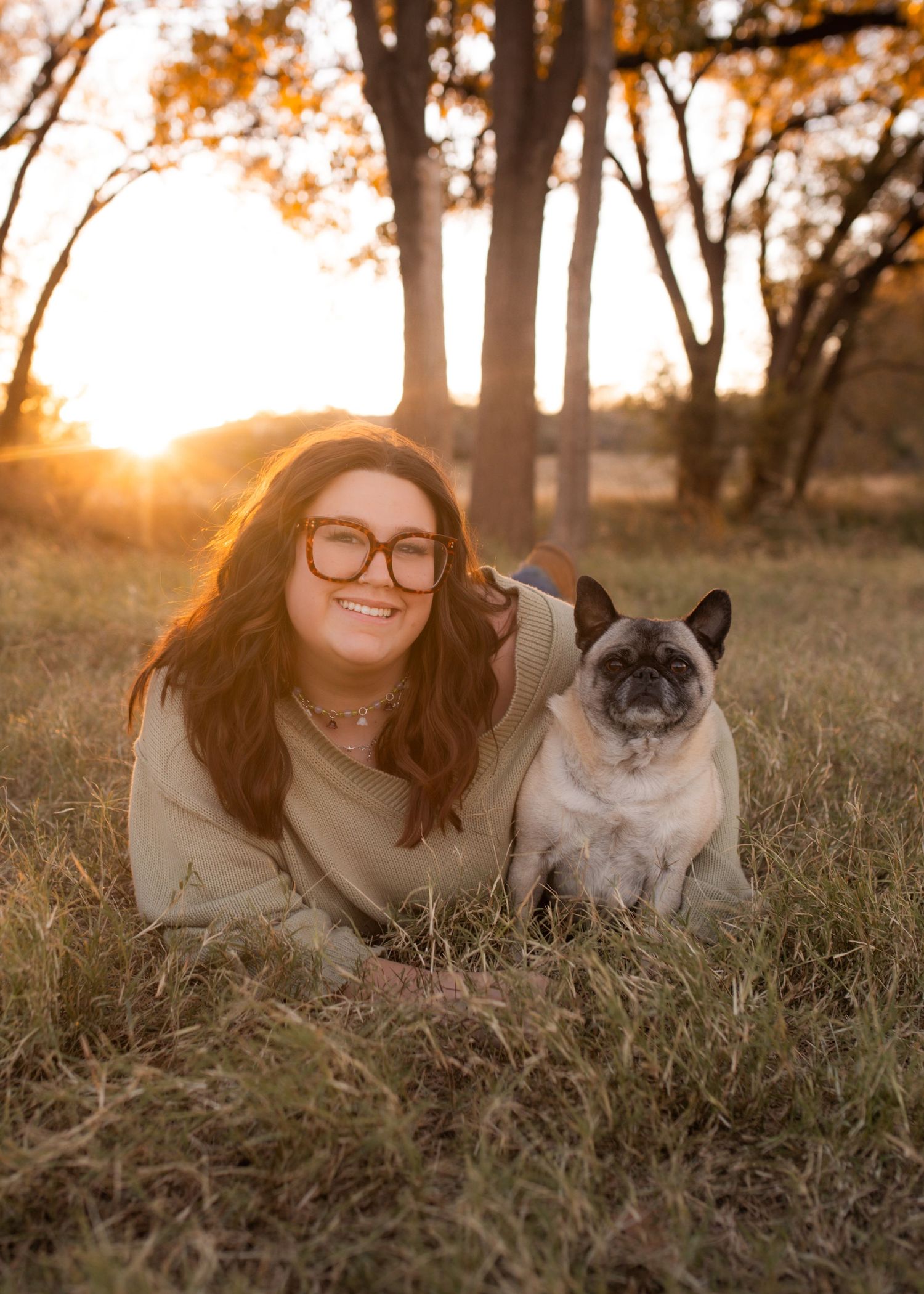 A smiling woman with glasses lies in golden grass at sunset beside a fawn French Bulldog in a scenic park.