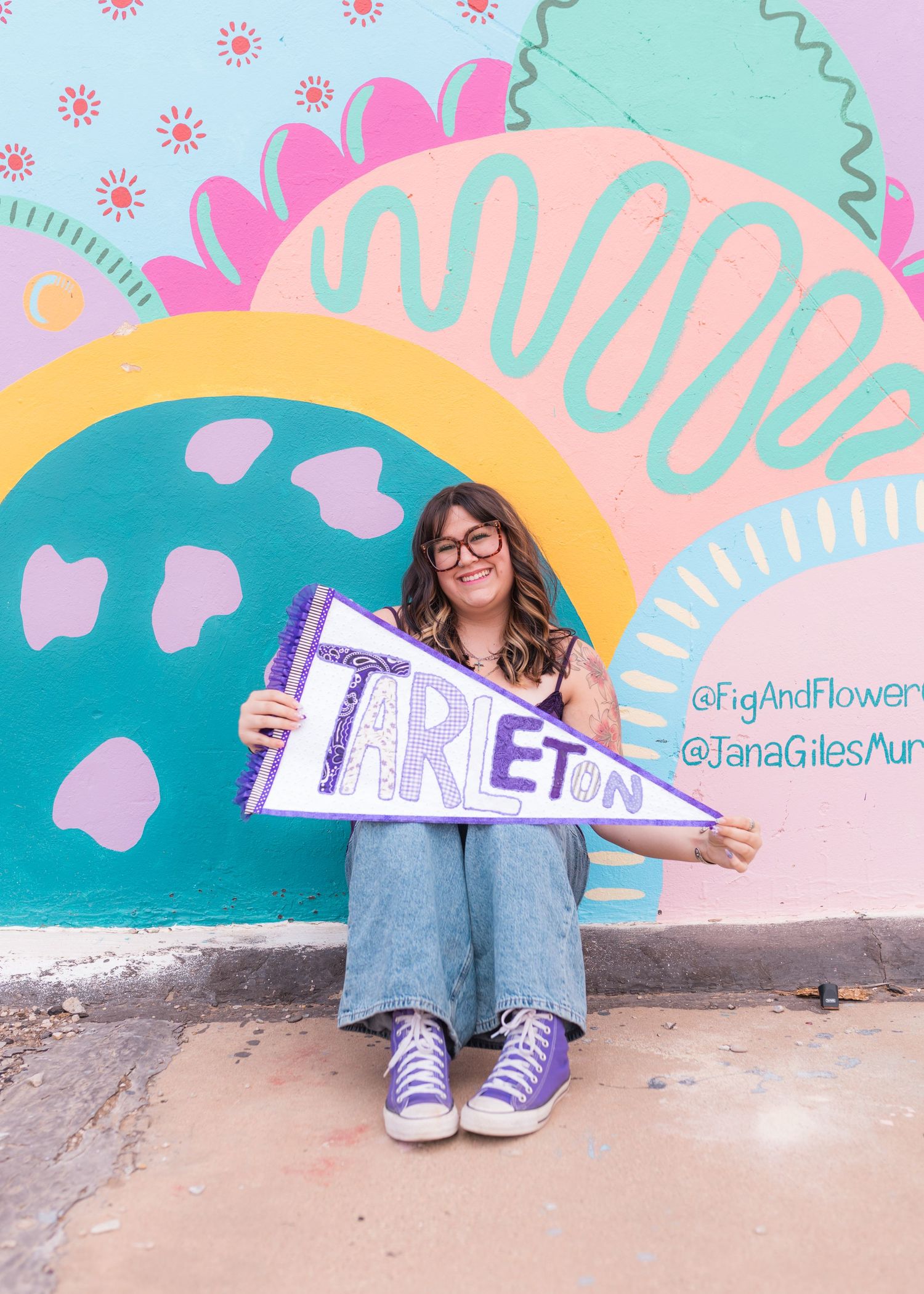 A young woman sits against a colorful mural holding a purple Tarleton pennant, wearing glasses and blue jeans.