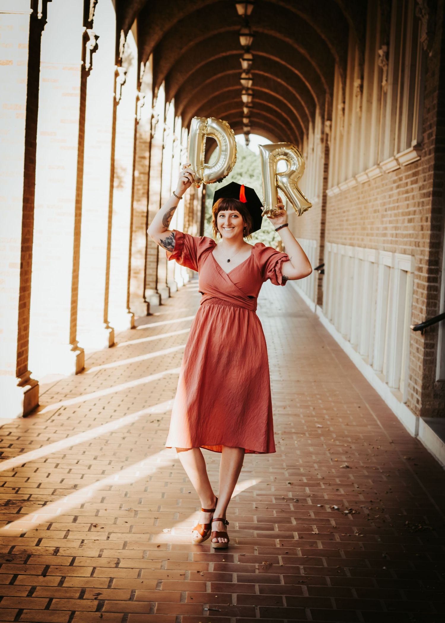 Person in red dress holding DR balloons in an arched hallway with dramatic lighting.