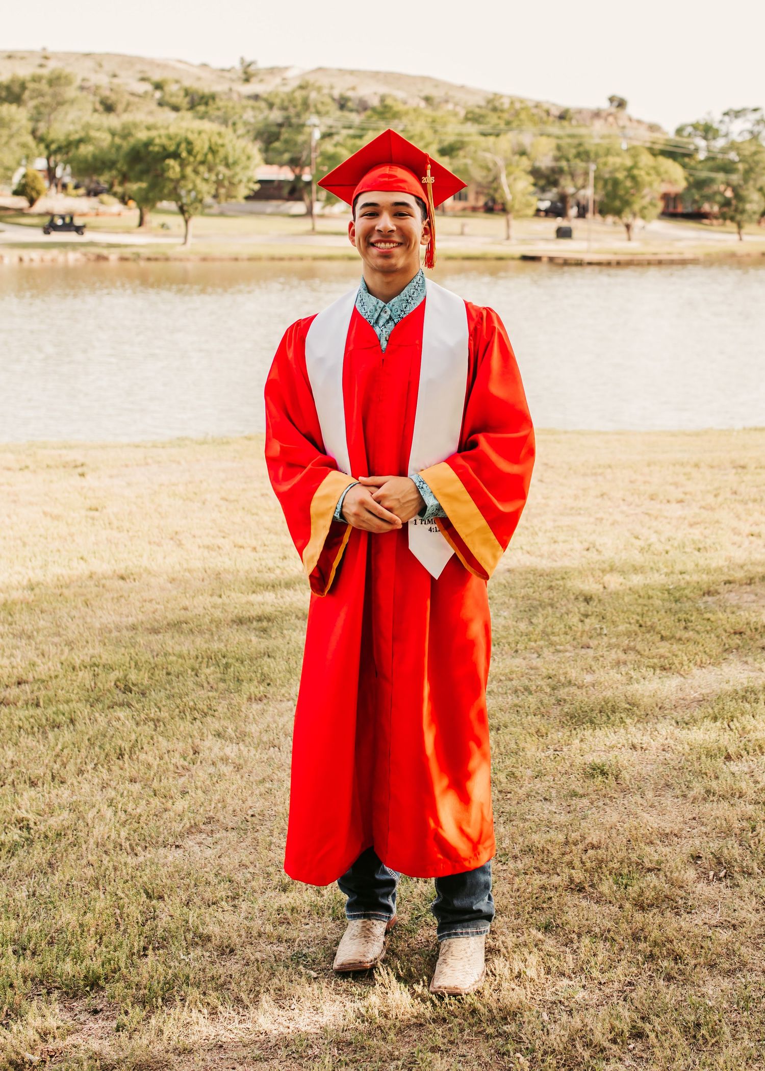 Graduate in red cap and gown smiling outdoors by lake.