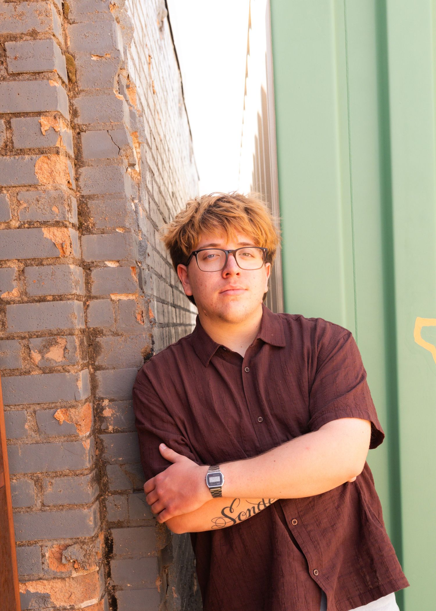 Young man with glasses and tattoo leans against brick wall in maroon shirt, arms crossed, outdoor urban setting.