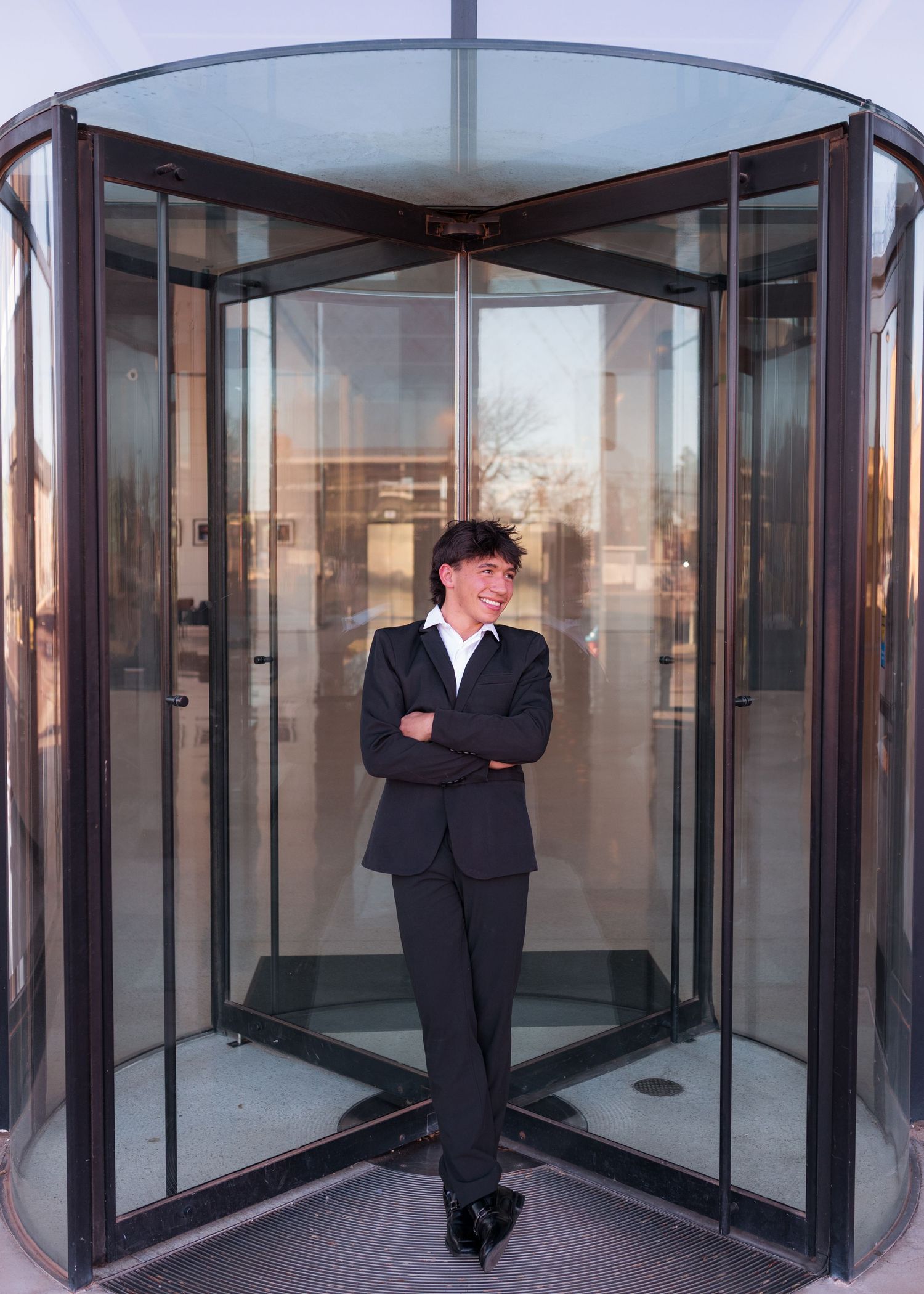 Young professional in black suit standing confidently with arms crossed in front of a glass revolving door entrance.