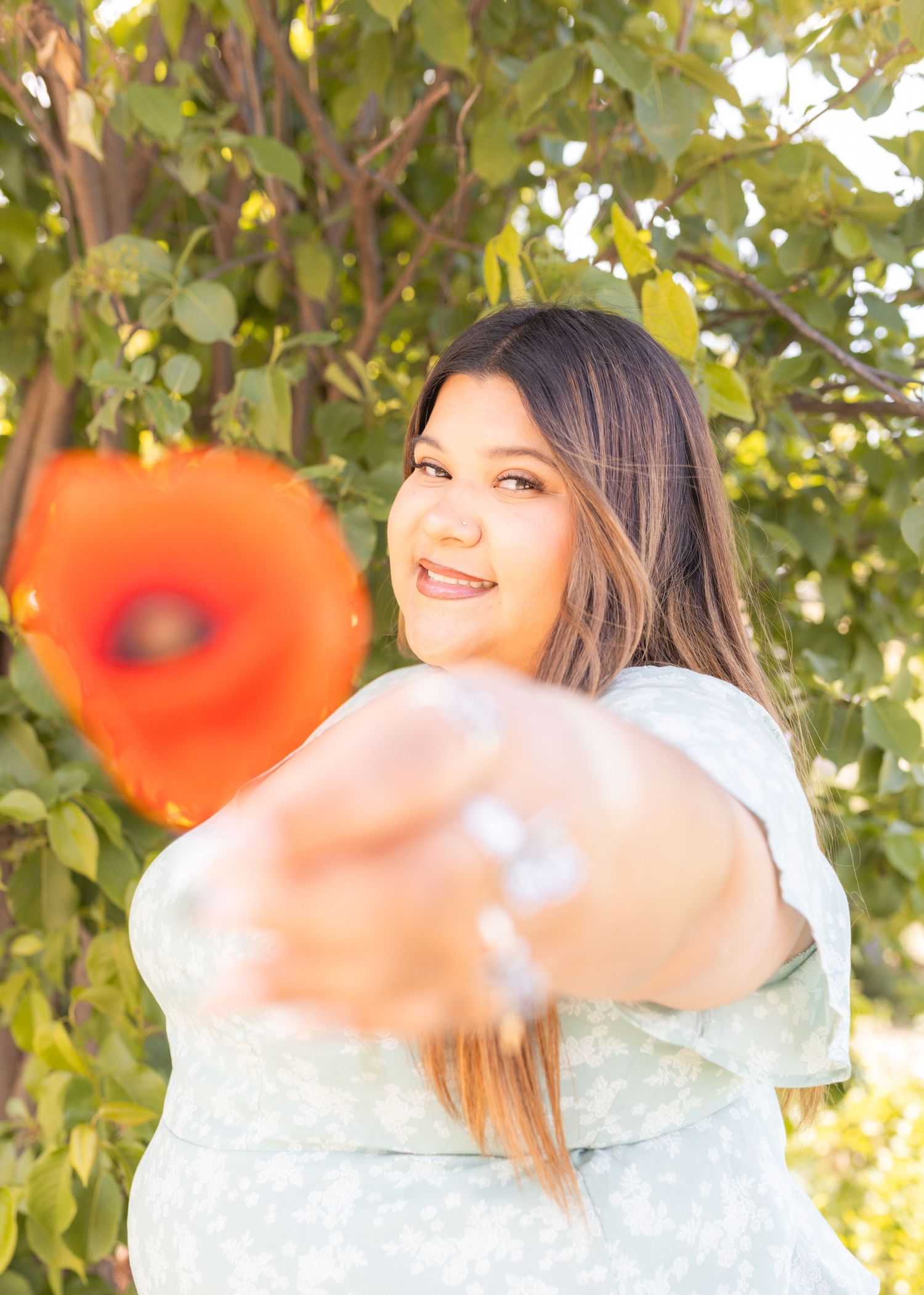 Young woman smiling outdoors holding a vibrant red poppy flower toward camera with green trees in background.