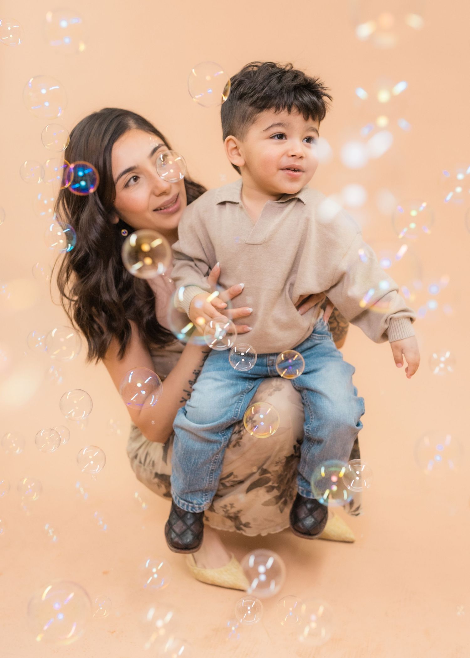 A young woman and toddler boy pose together surrounded by floating soap bubbles on a beige background.