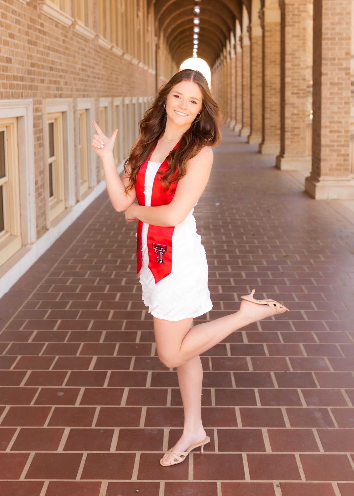 Texas Tech University graduate in white dress and red TTU stole poses playfully in brick archway hallway.