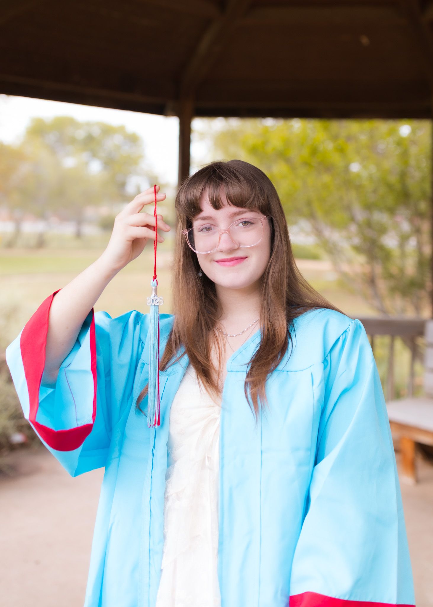 A graduate in light blue academic regalia holds a red tassel while standing under a covered outdoor area on graduation day.