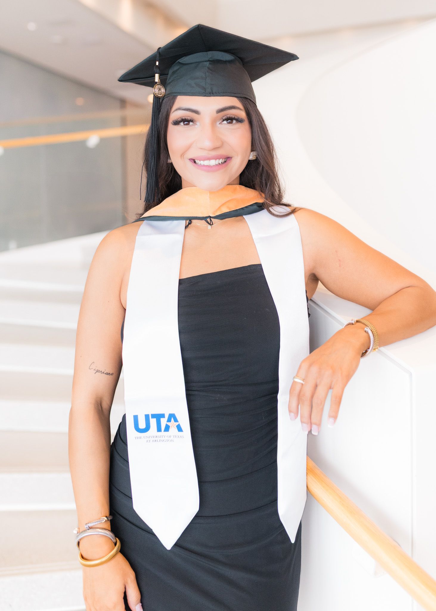 UTA graduate in cap and gown poses with white graduation stole in modern hallway.