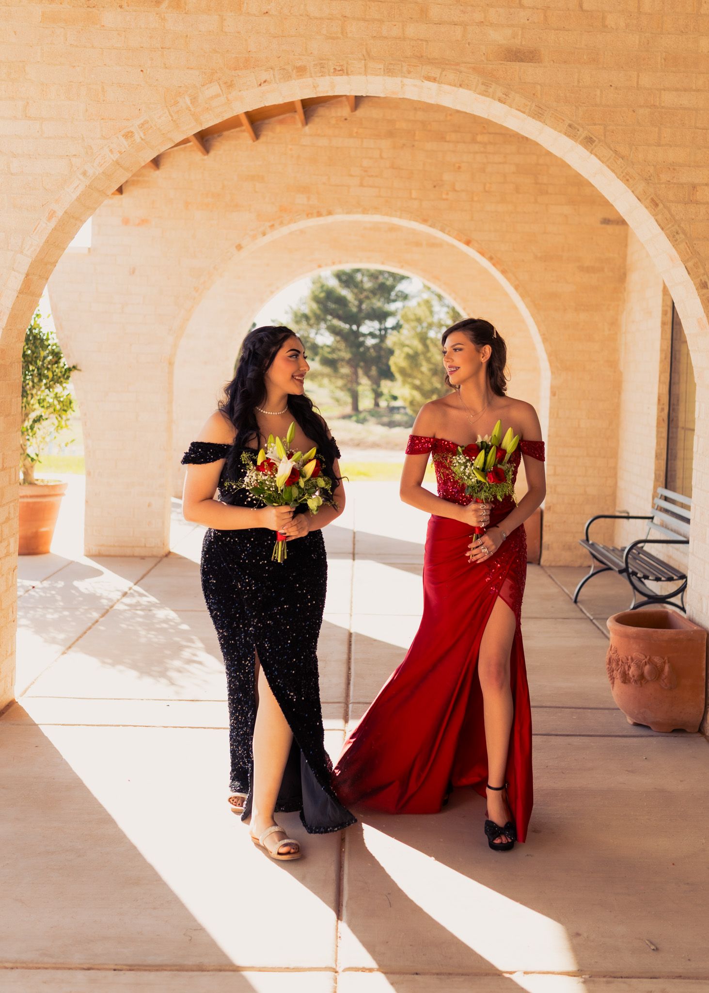 Two elegant women in formal dresses hold bouquets under arched walkway.