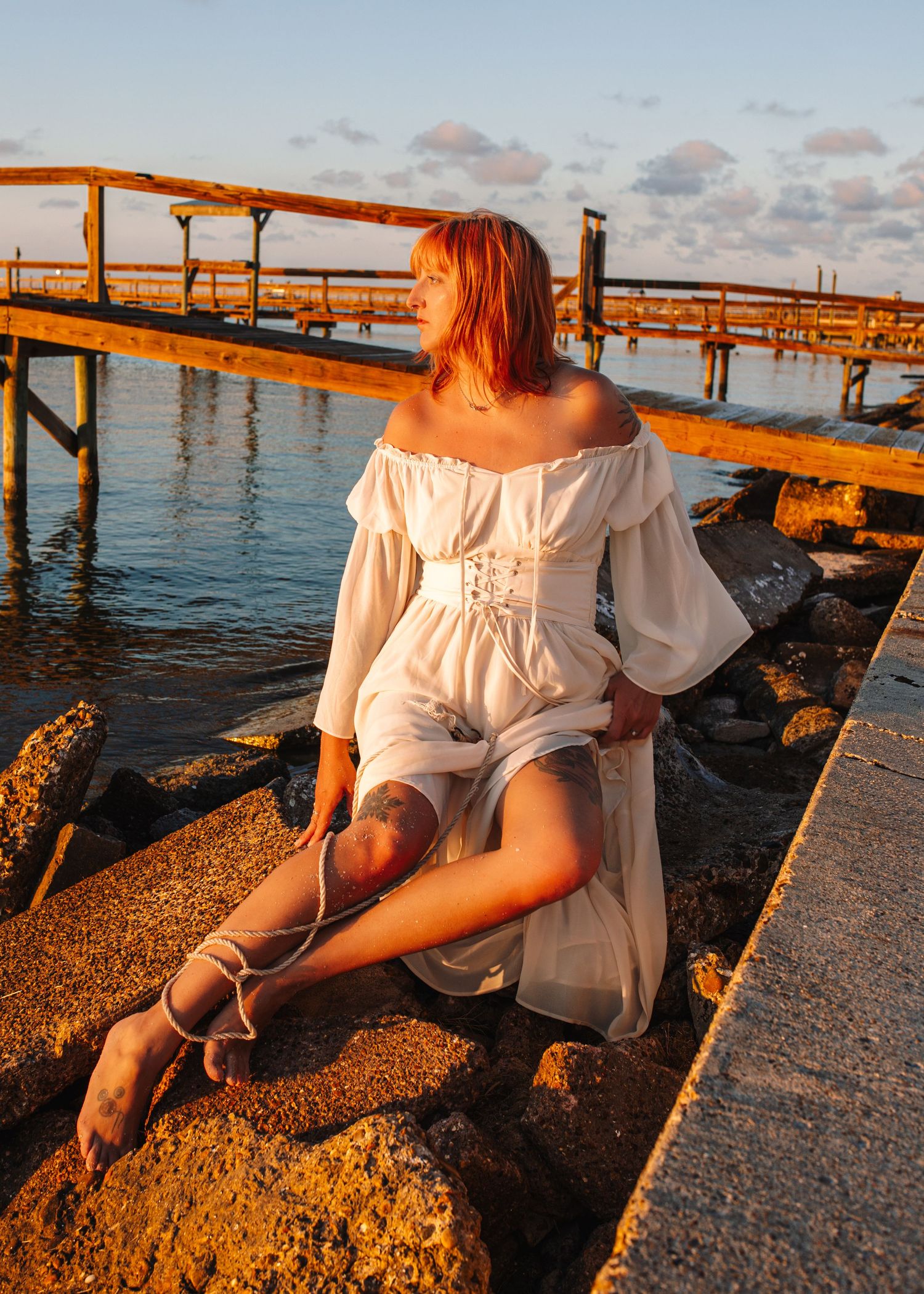 Woman in white off-shoulder dress sitting on rocks by a pier at golden hour sunset near the ocean.