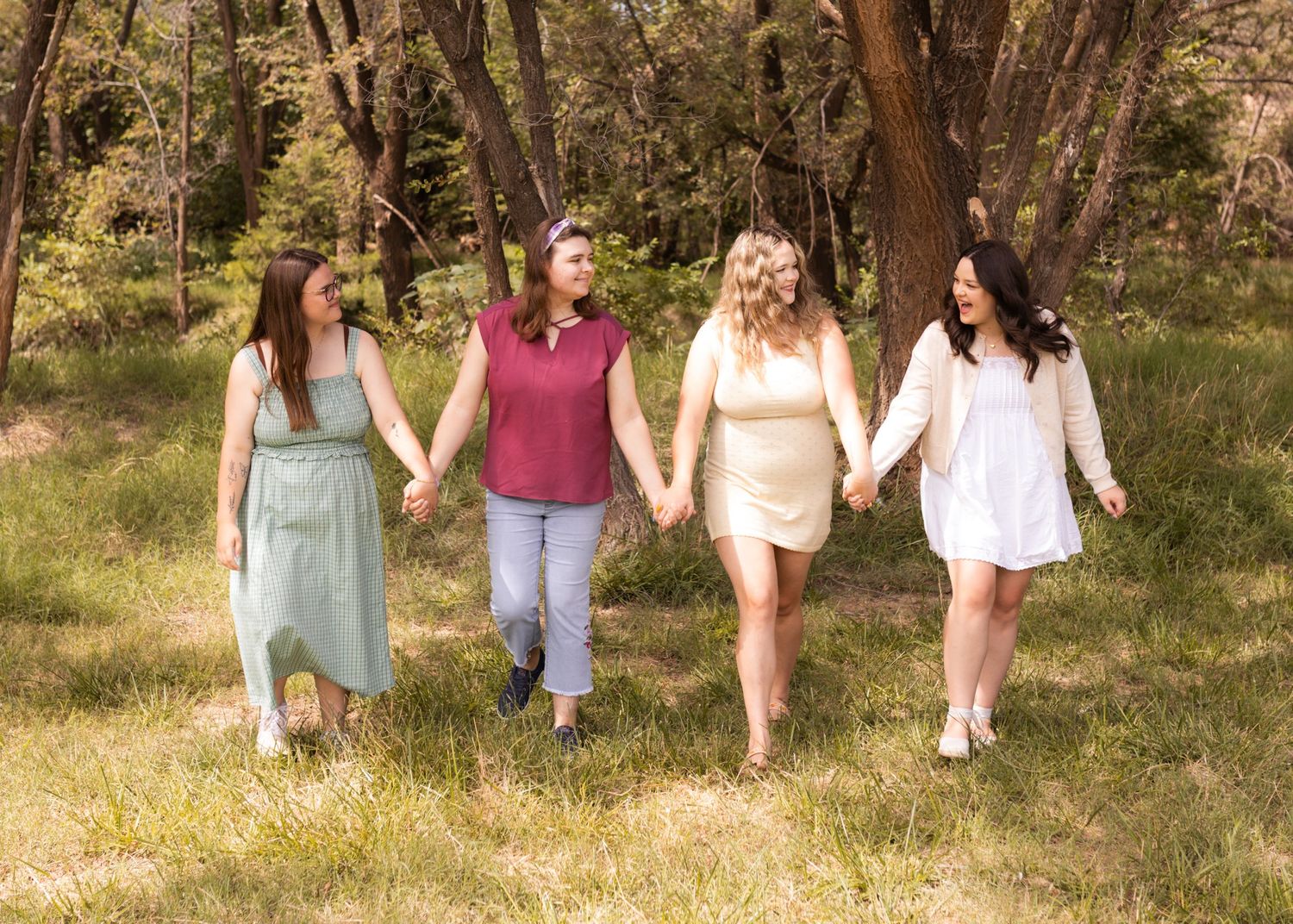 Four women holding hands walking through a sunny green meadow with trees in the background.