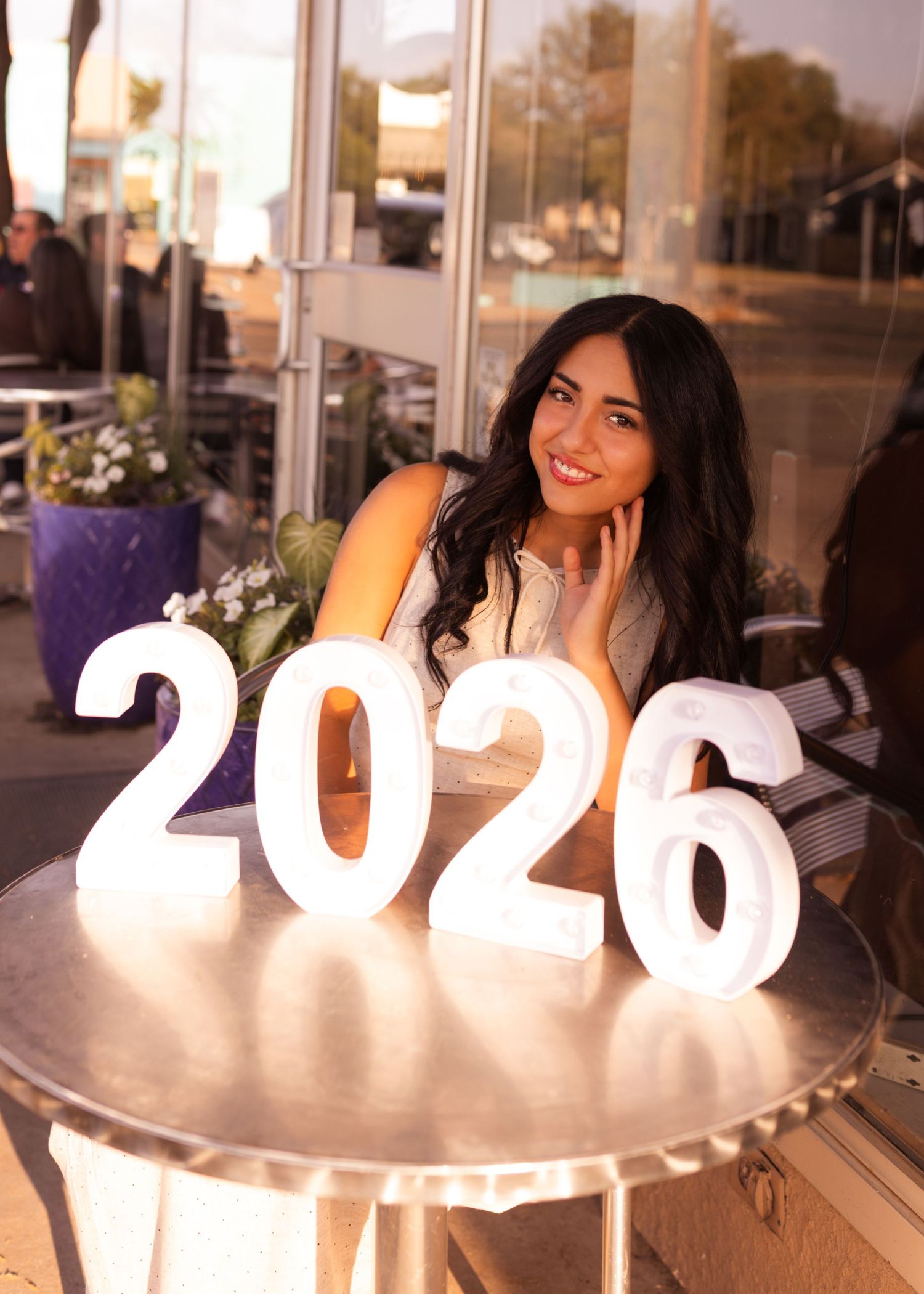 Young woman smiling behind a glowing 2026 sign on a table, celebrating her upcoming graduation outdoors.