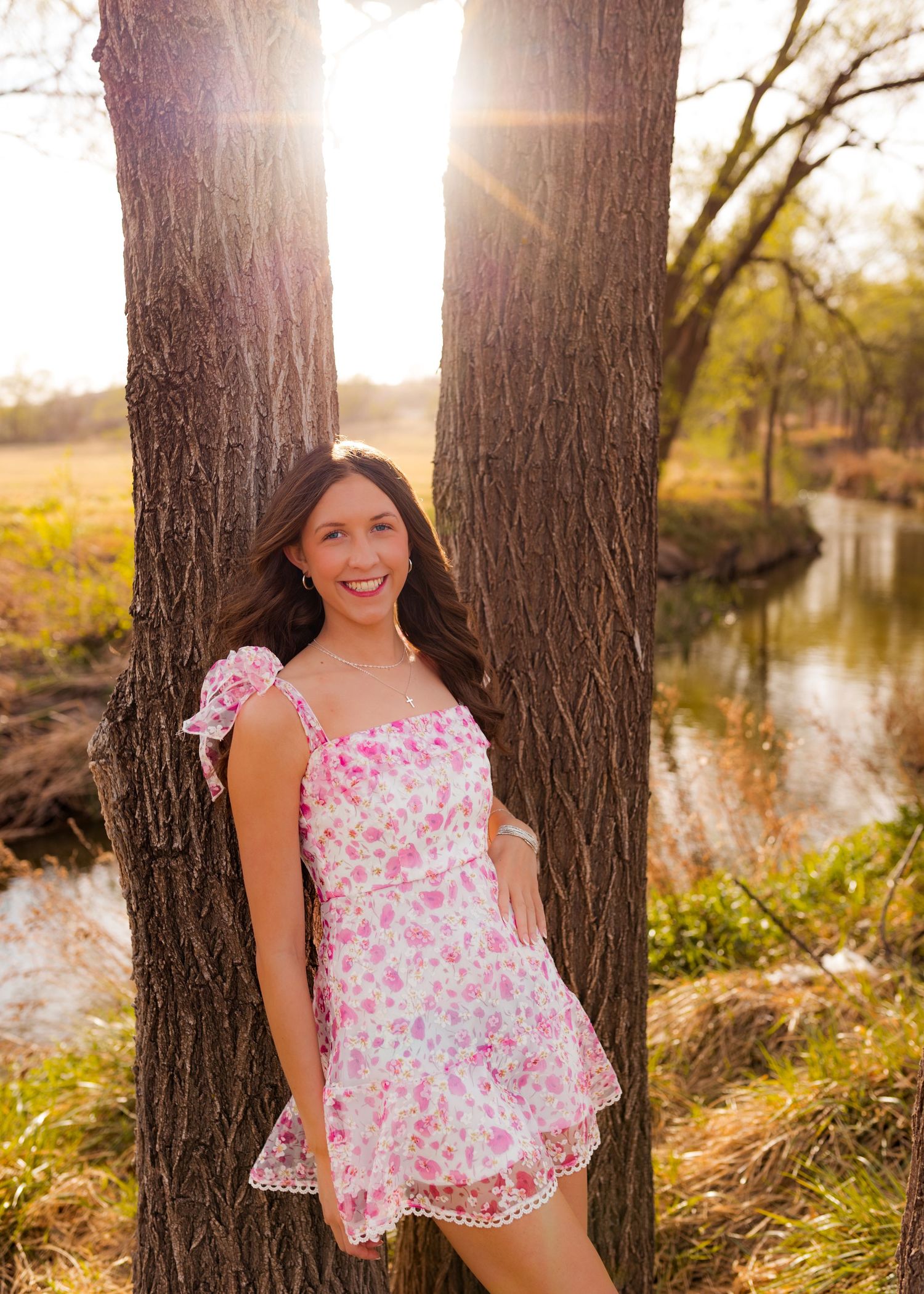 Young woman in pink floral dress poses between trees at golden hour near a serene pond.