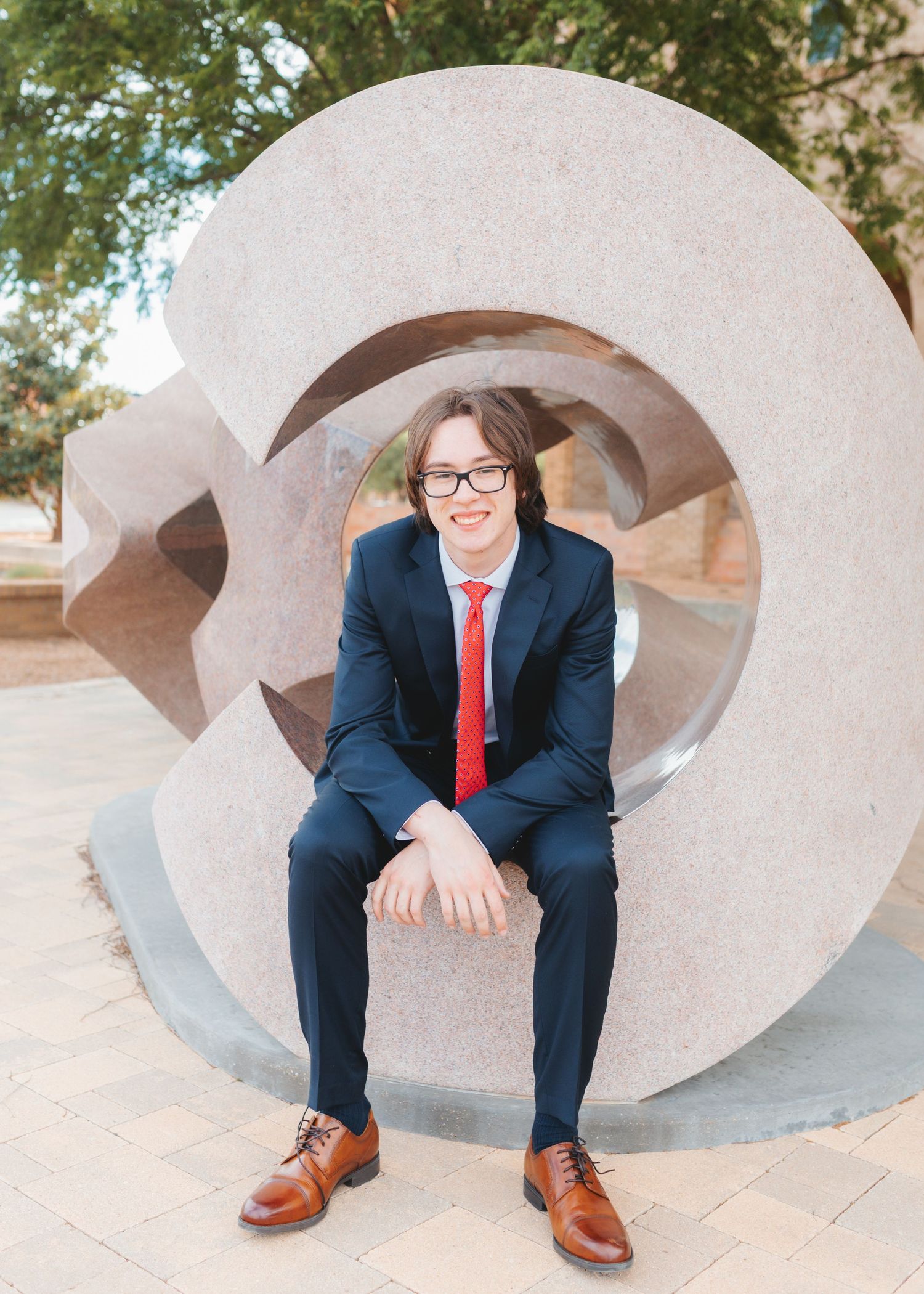 Young man in navy suit and red tie sitting on a large circular stone sculpture outdoors.
