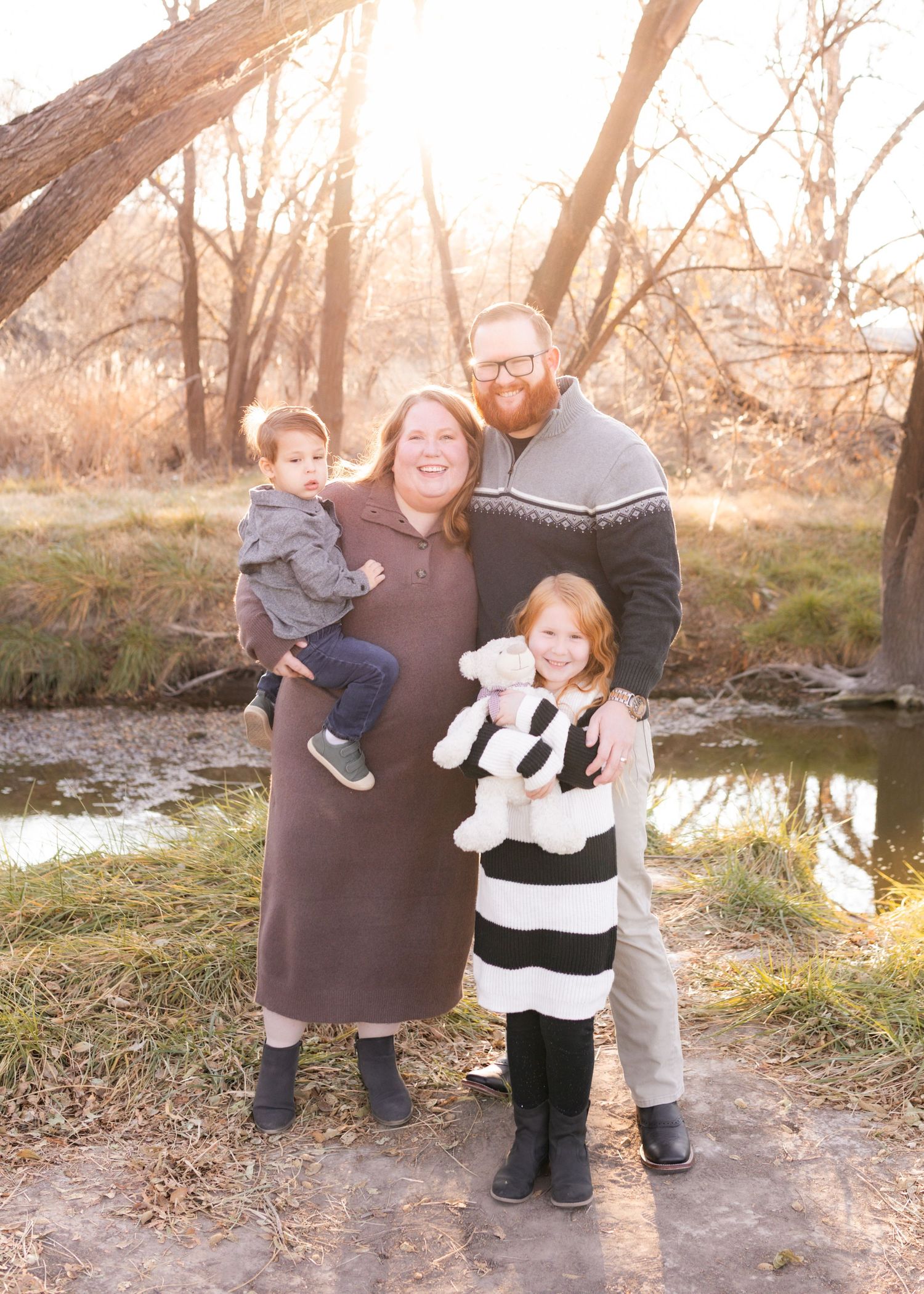 Family of four posing outdoors near a creek in autumn, parents holding children, golden hour lighting among bare trees.