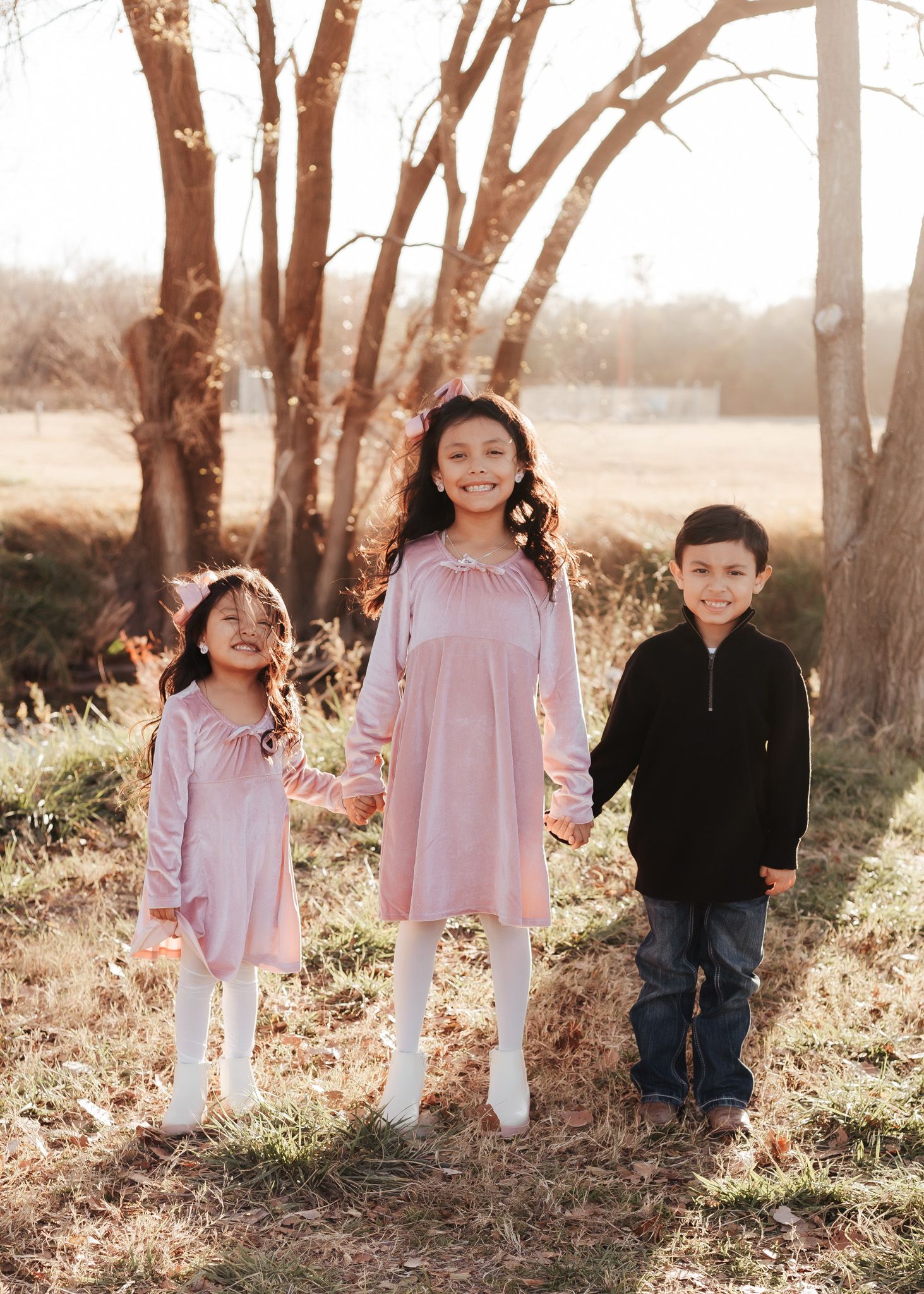 Three children in coordinated outfits standing together in a sunlit wooded area during golden hour.