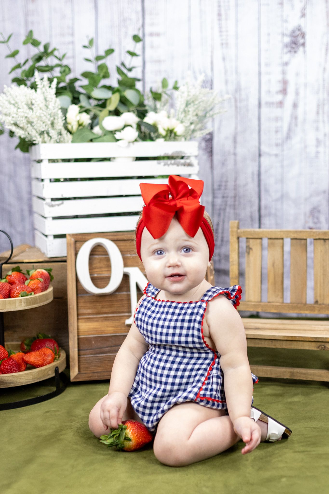 Baby in gingham romper with red bow poses with strawberries and rustic decor on green backdrop.