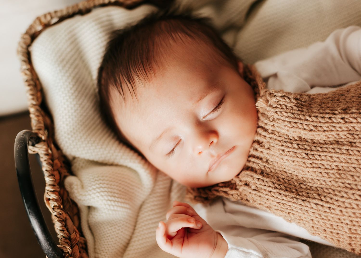 A sleeping baby swaddled in cream knit blankets rests peacefully in a woven basket.