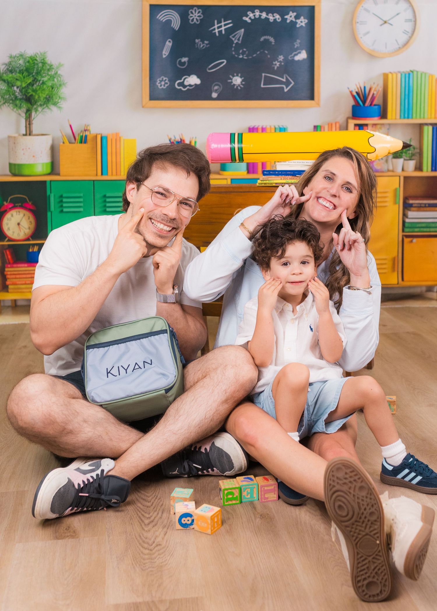 Family of three sitting on floor in colorful classroom, smiling and posing with Kiyan bag, with chalkboard and toys.